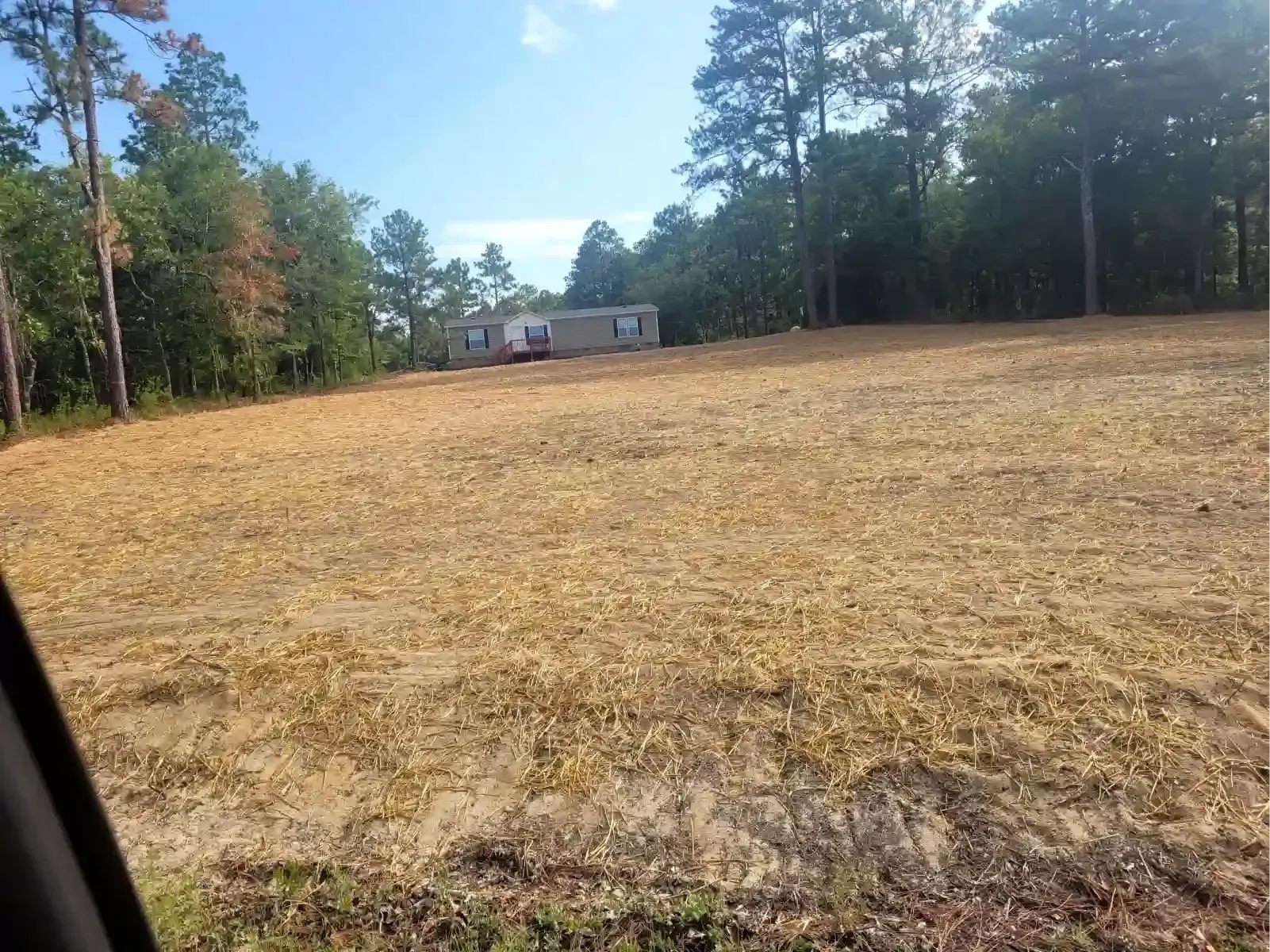 A wide, cleared plot of land with dry grass slopes up toward a distant manufactured home surrounded by trees.