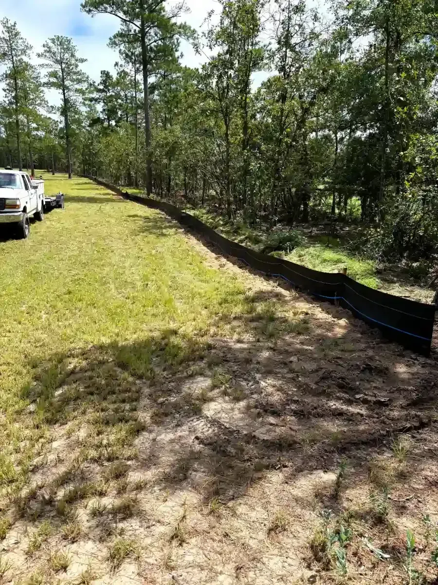 A black silt fence runs along the edge of a grassy field bordered by a dense forest, with a white truck parked nearby.