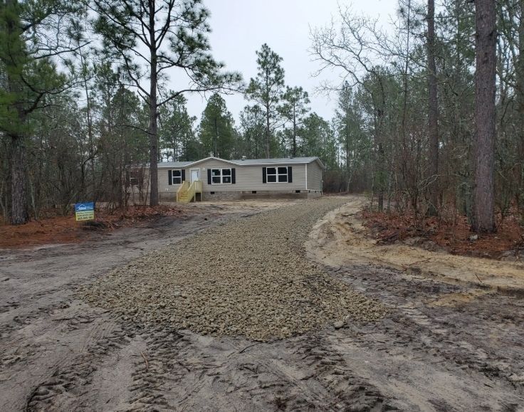 A manufactured home sits in a wooded lot at the end of a long, partially graveled dirt driveway.