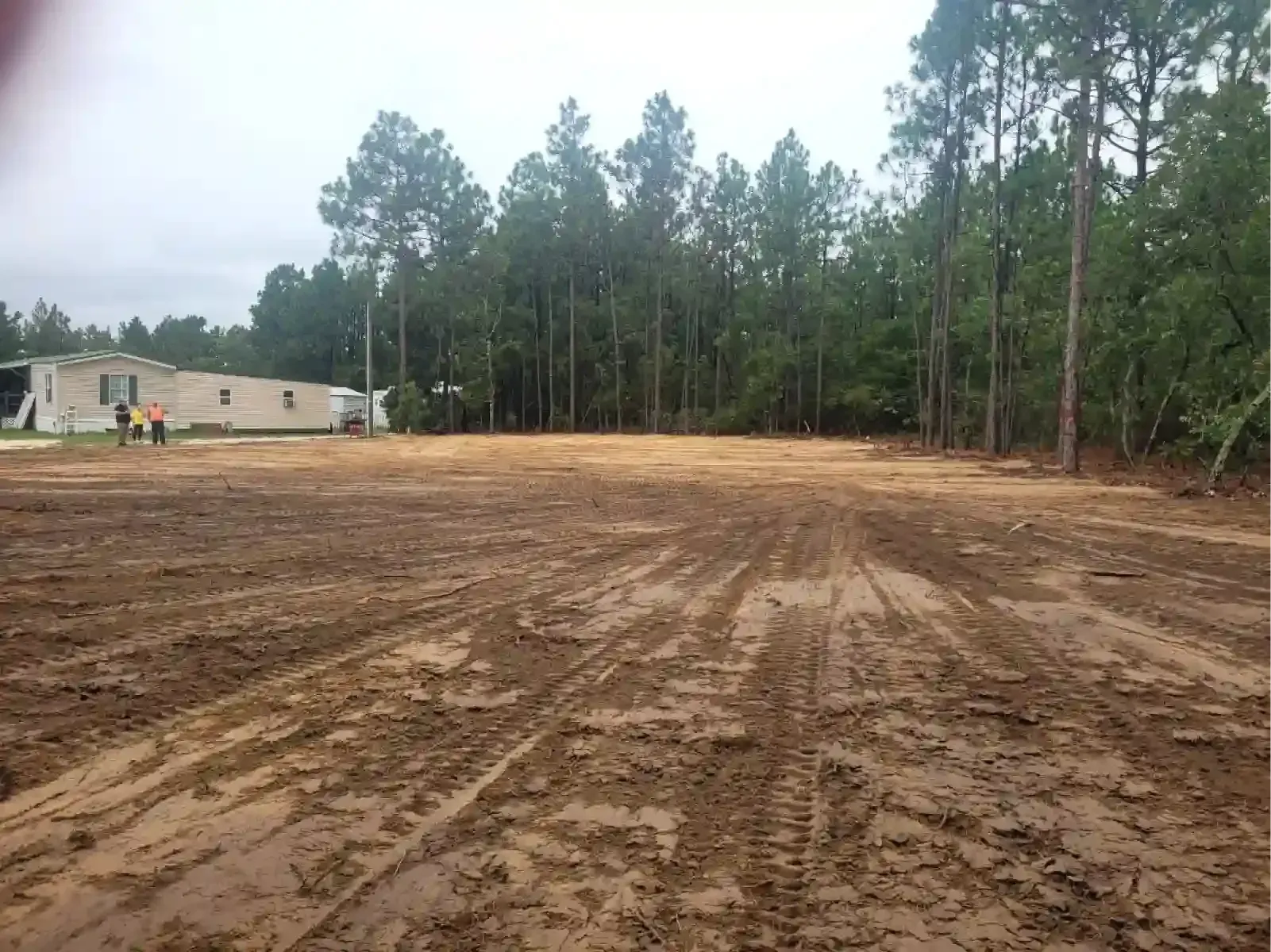 A cleared dirt lot with tire tracks in the foreground and a mobile home near a forest in the background.