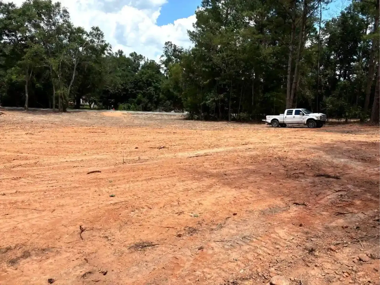A white pickup truck parked on a cleared, red-clay dirt lot next to a line of dense green trees under a blue sky.
