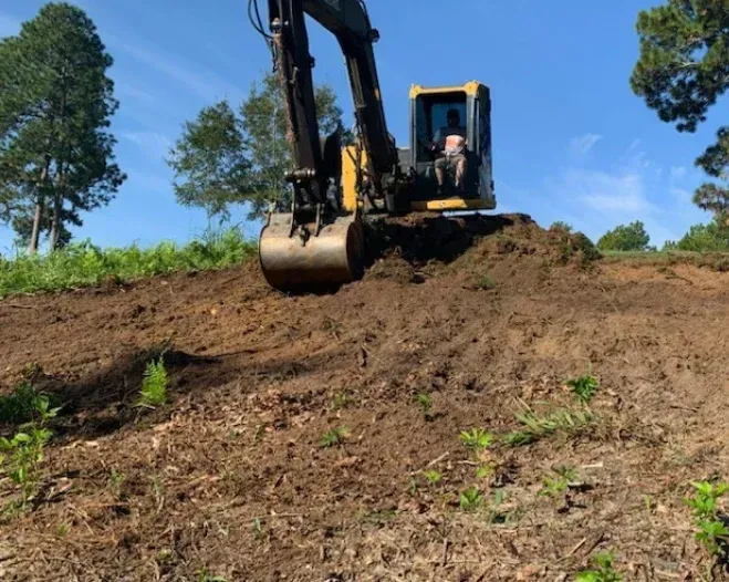 A yellow excavator moves dirt on a hillside under a clear blue sky.