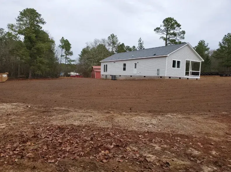 A white house with a screened porch sits on a cleared, dirt-covered lot near pine trees under a cloudy sky.