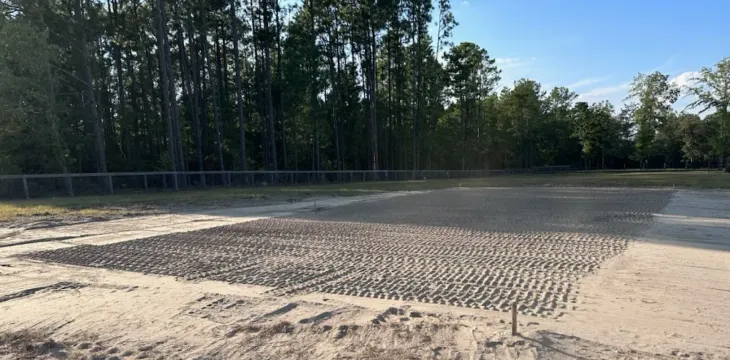 A rectangular area of fresh, textured earth or gravel surrounded by sandy ground and a line of trees under a blue sky.