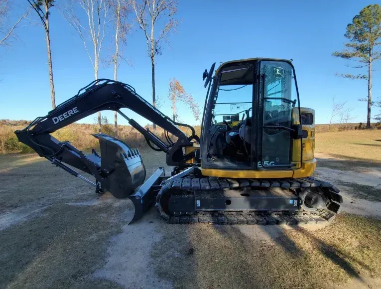 A yellow and black John Deere mini excavator parked on a dirt path in a grassy field under a clear blue sky.
