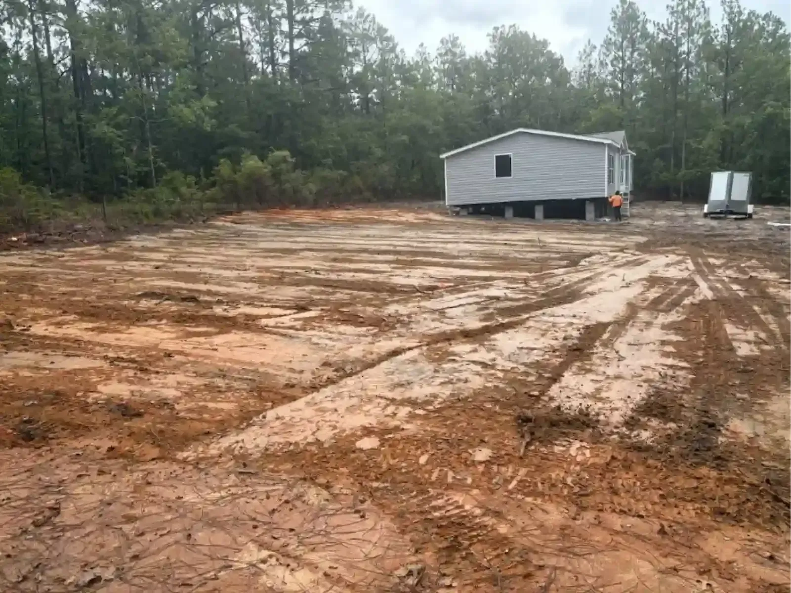 A leveled, muddy construction site with tire tracks leading toward a small, gray modular building in a wooded area.