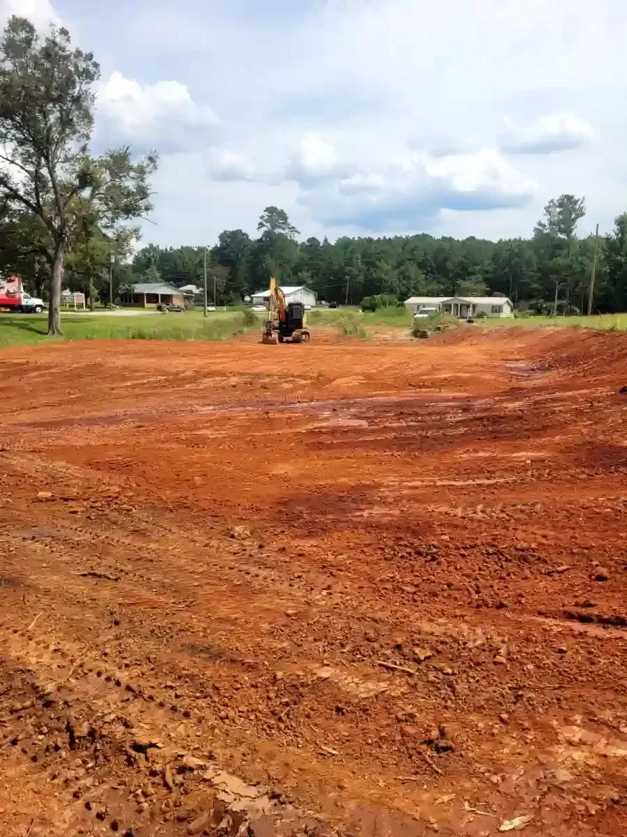 A construction excavator works on a cleared, uneven red clay lot with trees and houses in the distance under a blue sky.