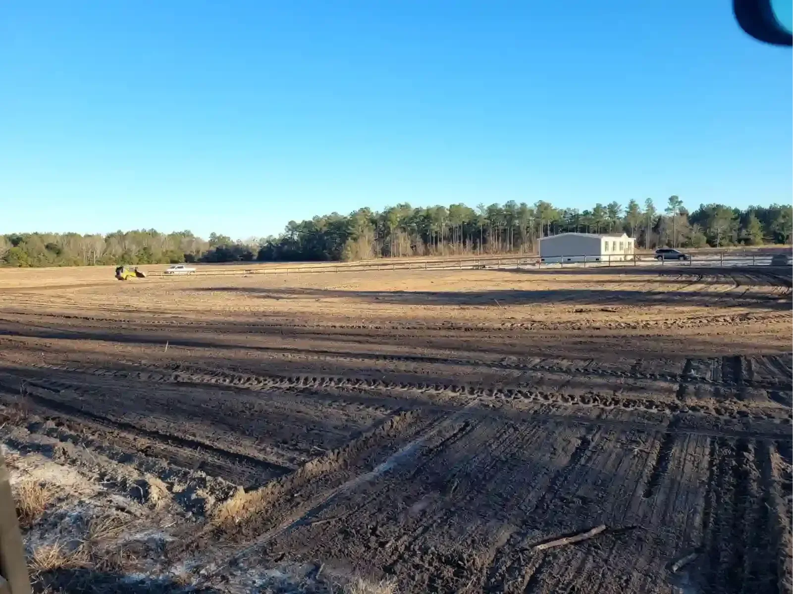 A wide, cleared construction site with dark soil, tire tracks in the foreground, and a small building under a blue sky.