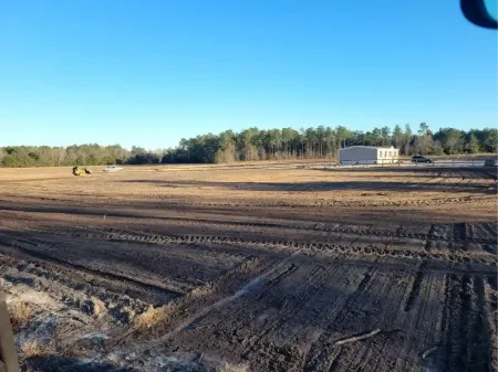 A wide, cleared construction site with dark soil, tire tracks in the foreground, and a small building under a blue sky.