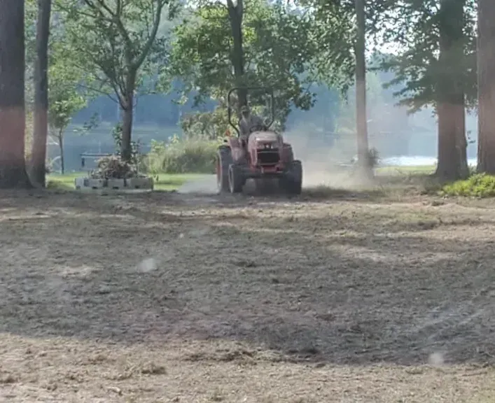 A red tractor kicks up dust while driving through a grassy, wooded area.