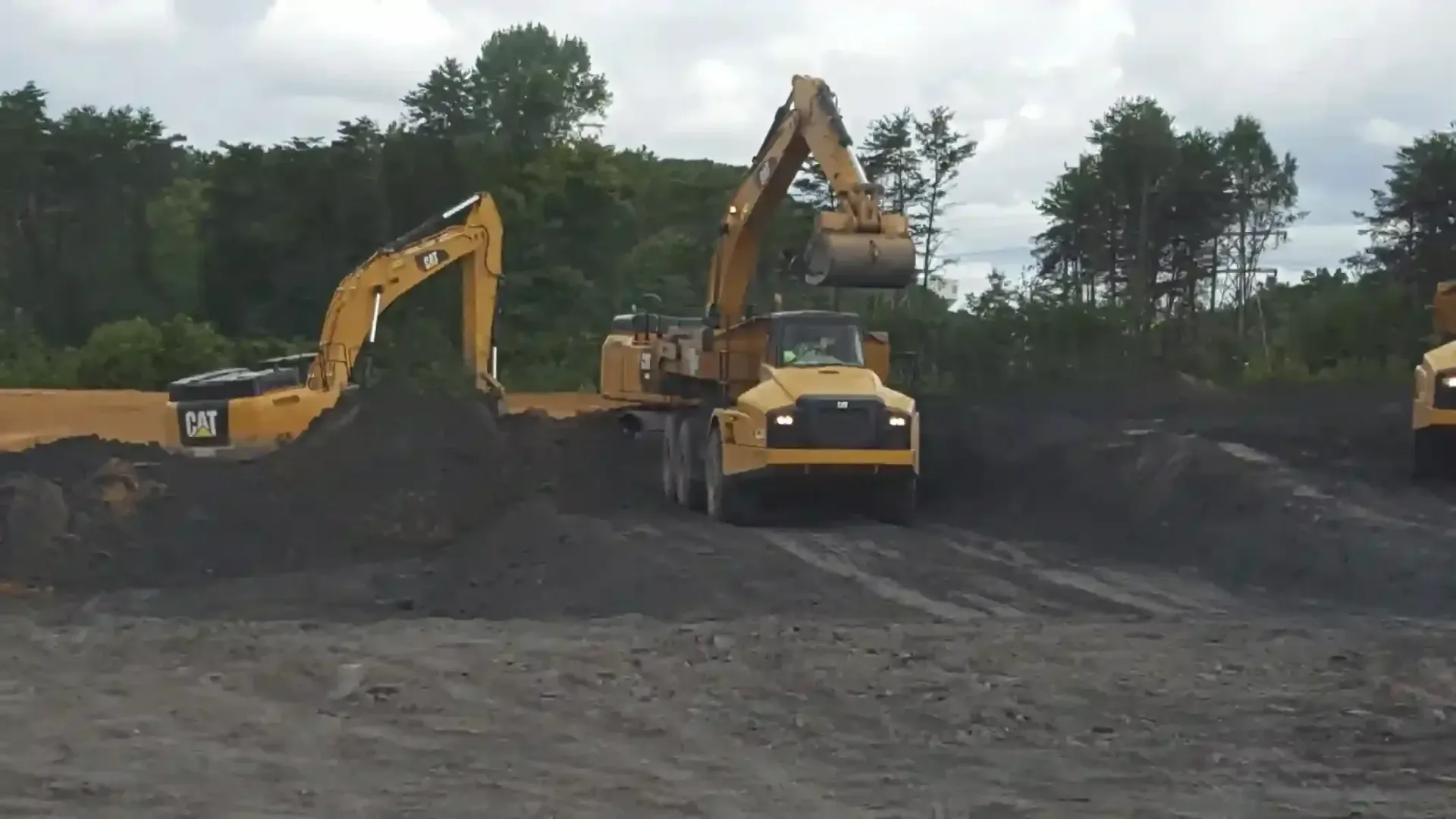 Two yellow CAT excavators operate in a dark soil pit under a cloudy sky.