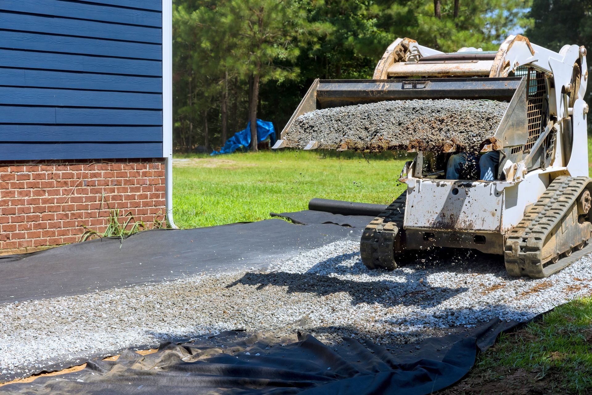 A white skid steer with a bucket of gravel drives over landscape fabric next to a blue-sided house with a brick foundation.