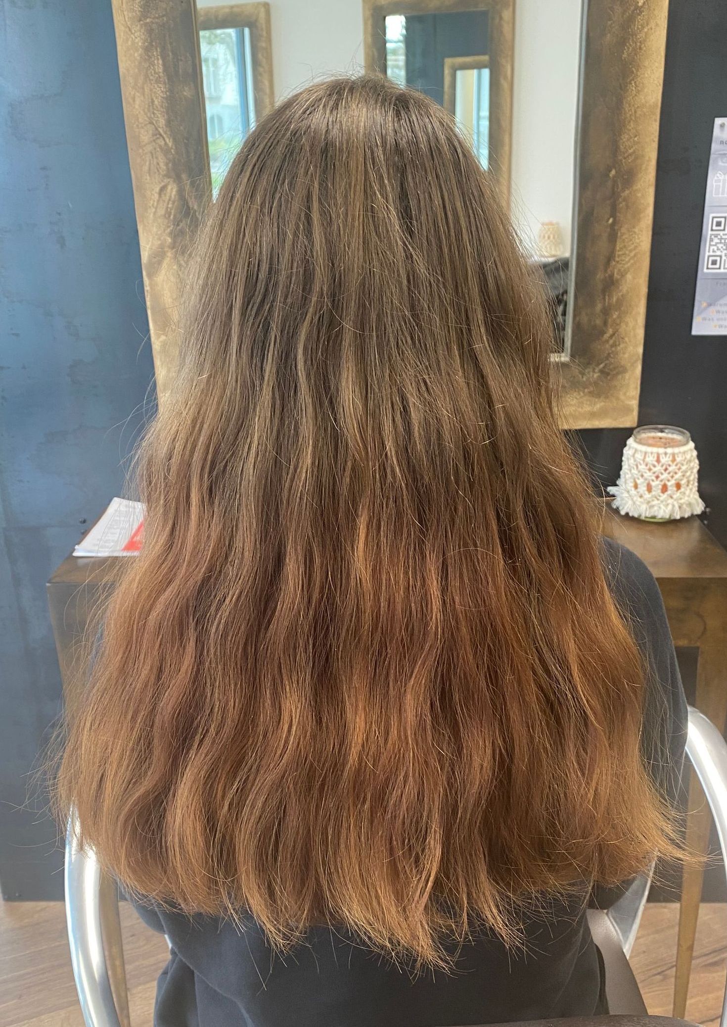 Rückansicht einer Frau mit langem, lockigem Haar. Das Haar ist eine Mischung aus Braun- und Kupfertönen. Sitzt in einem Salon.