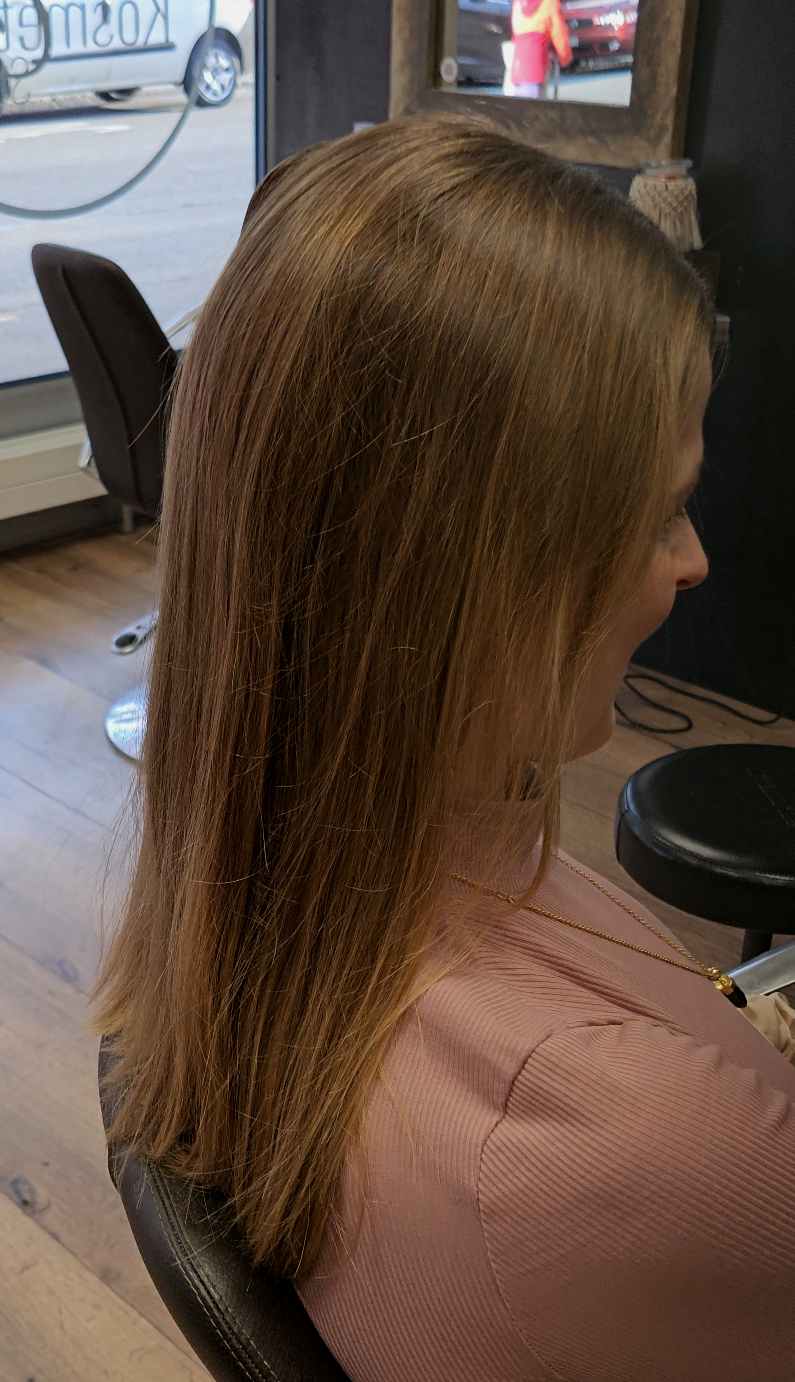 Frau mit schulterlangem braunem Haar sitzt in einem Friseurstuhl. Sie trägt einen rosa Pullover.