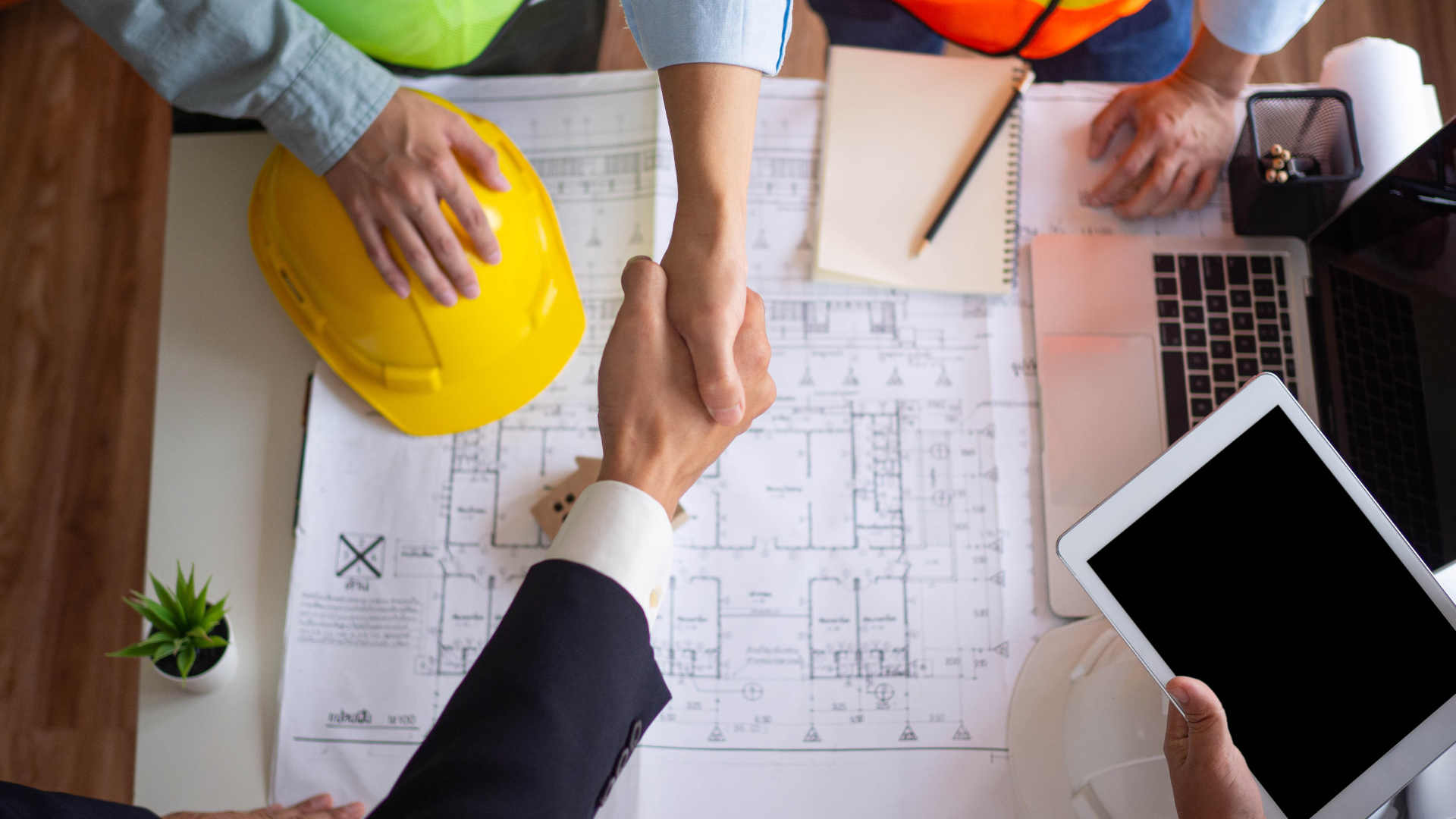 A high-angle view of business partners shaking hands over building blueprints, with a tablet, laptop, and hard hat nearby.