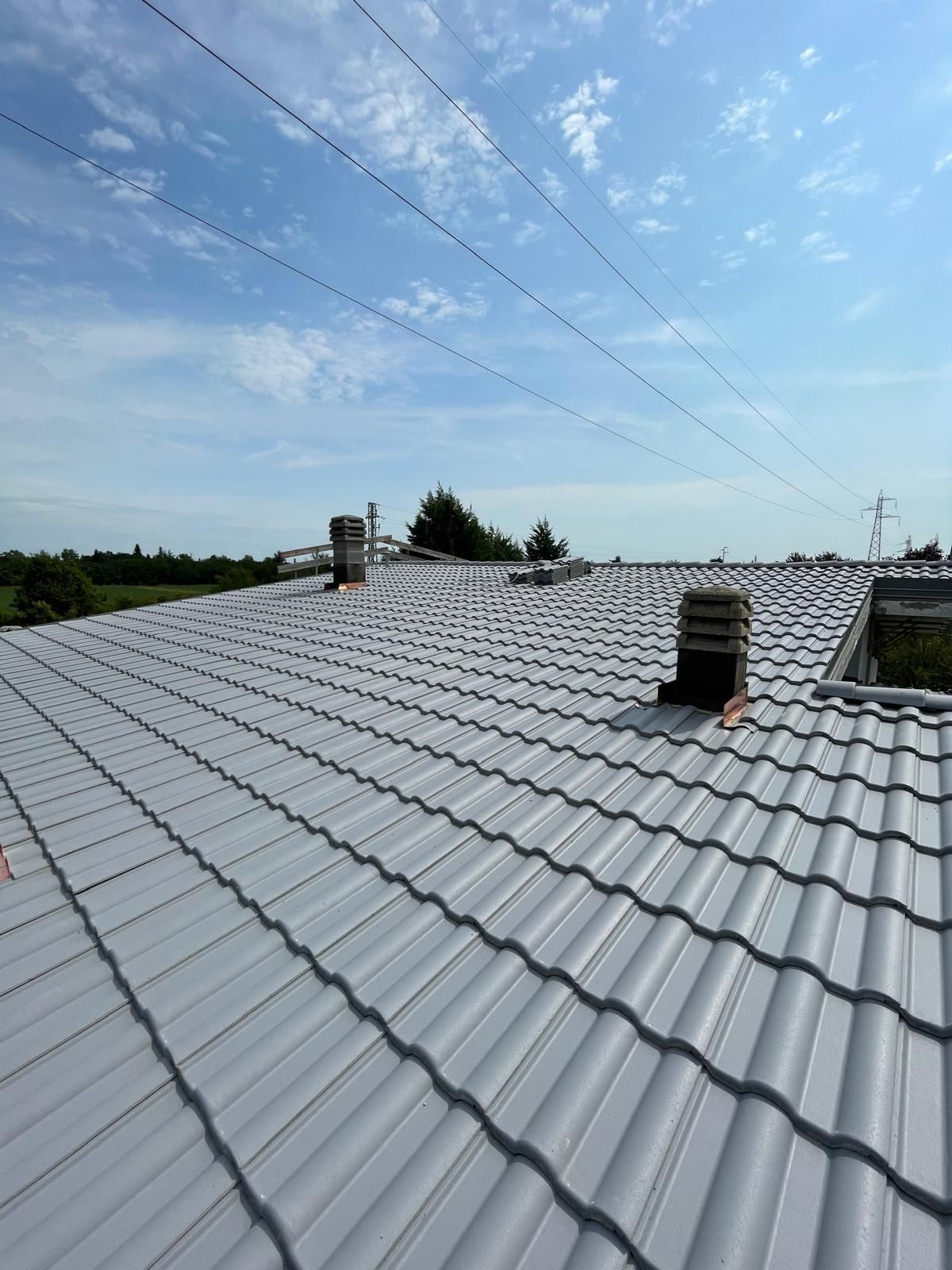Tetto in metallo grigio con tre camini sotto un cielo azzurro, linee elettriche in alto, campi sullo sfondo.