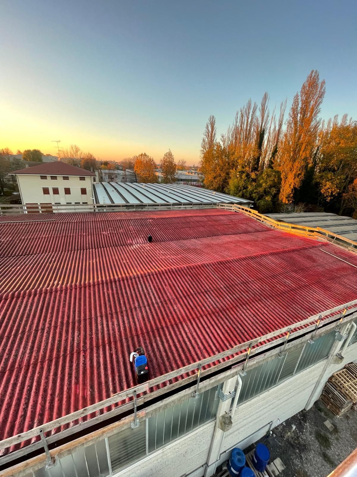 Tetto rosso ondulato con una persona che lavora; alberi ed edifici sullo sfondo sotto un cielo blu e arancione.