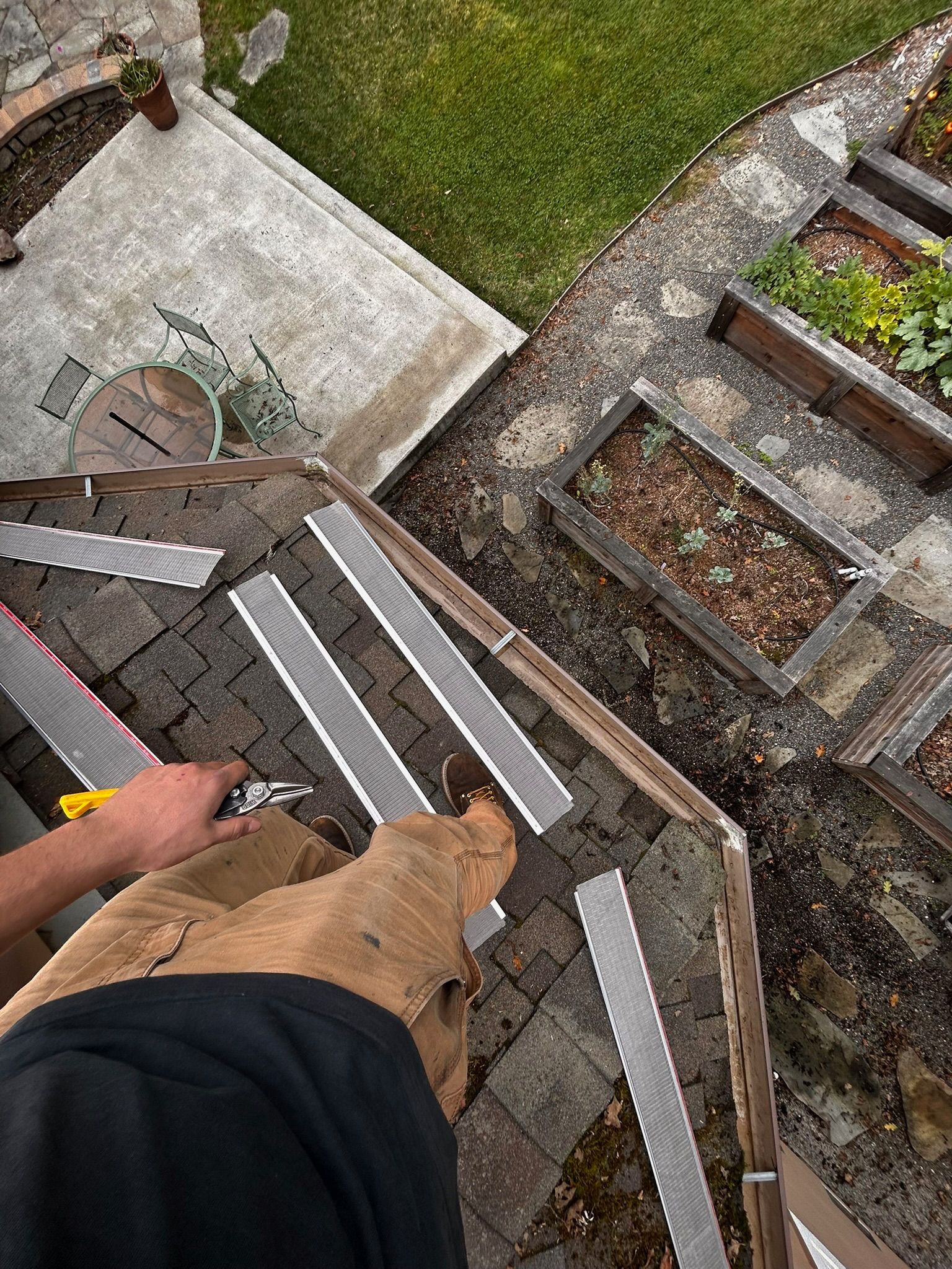Person on a roof installing metal flashing, using tools on a brown shingled roof with a garden in the background.
