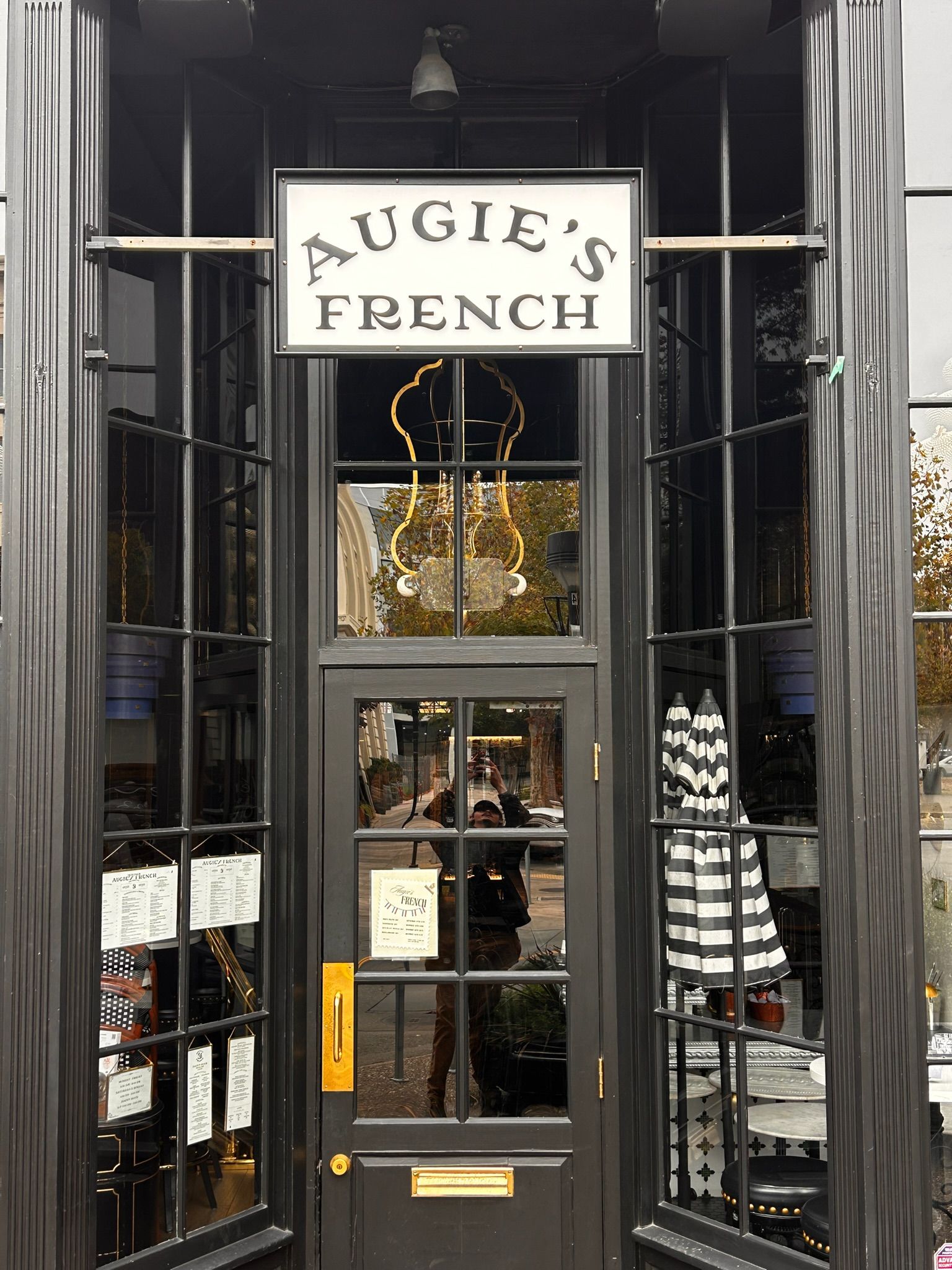 Exterior of Augie's French restaurant with black framed windows and door. Sign over door. Striped umbrella visible.