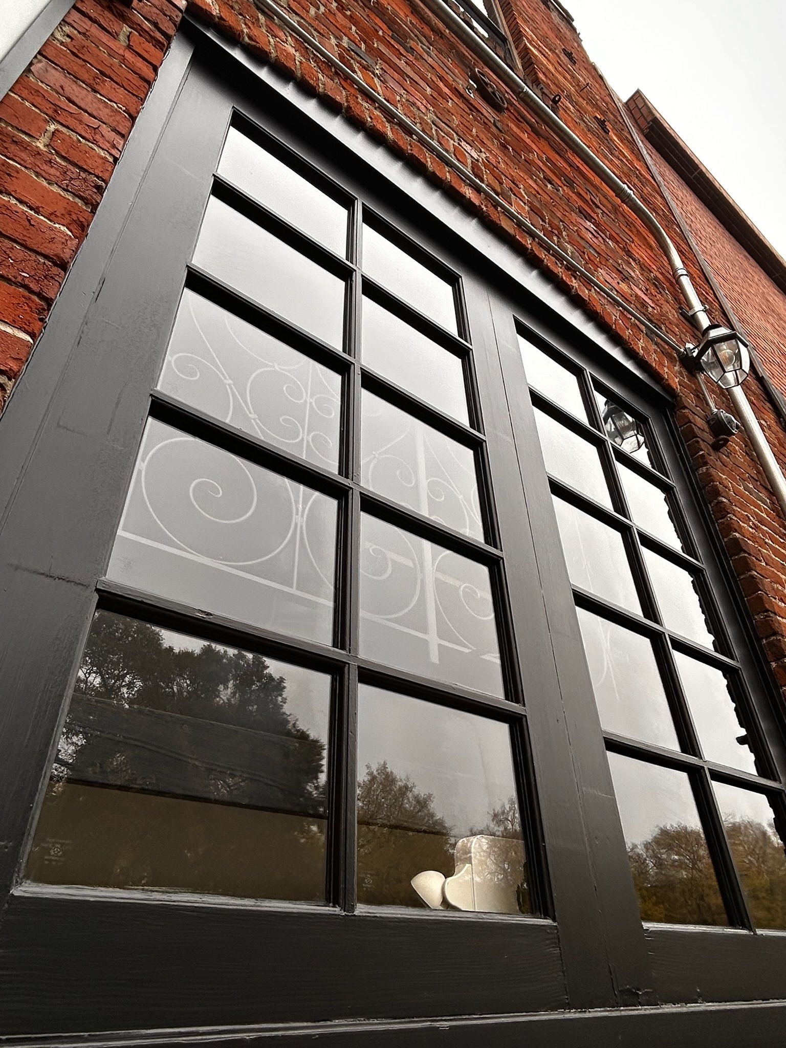 Black-framed multi-pane window in red brick building; window reflects a tree and light fixtures.