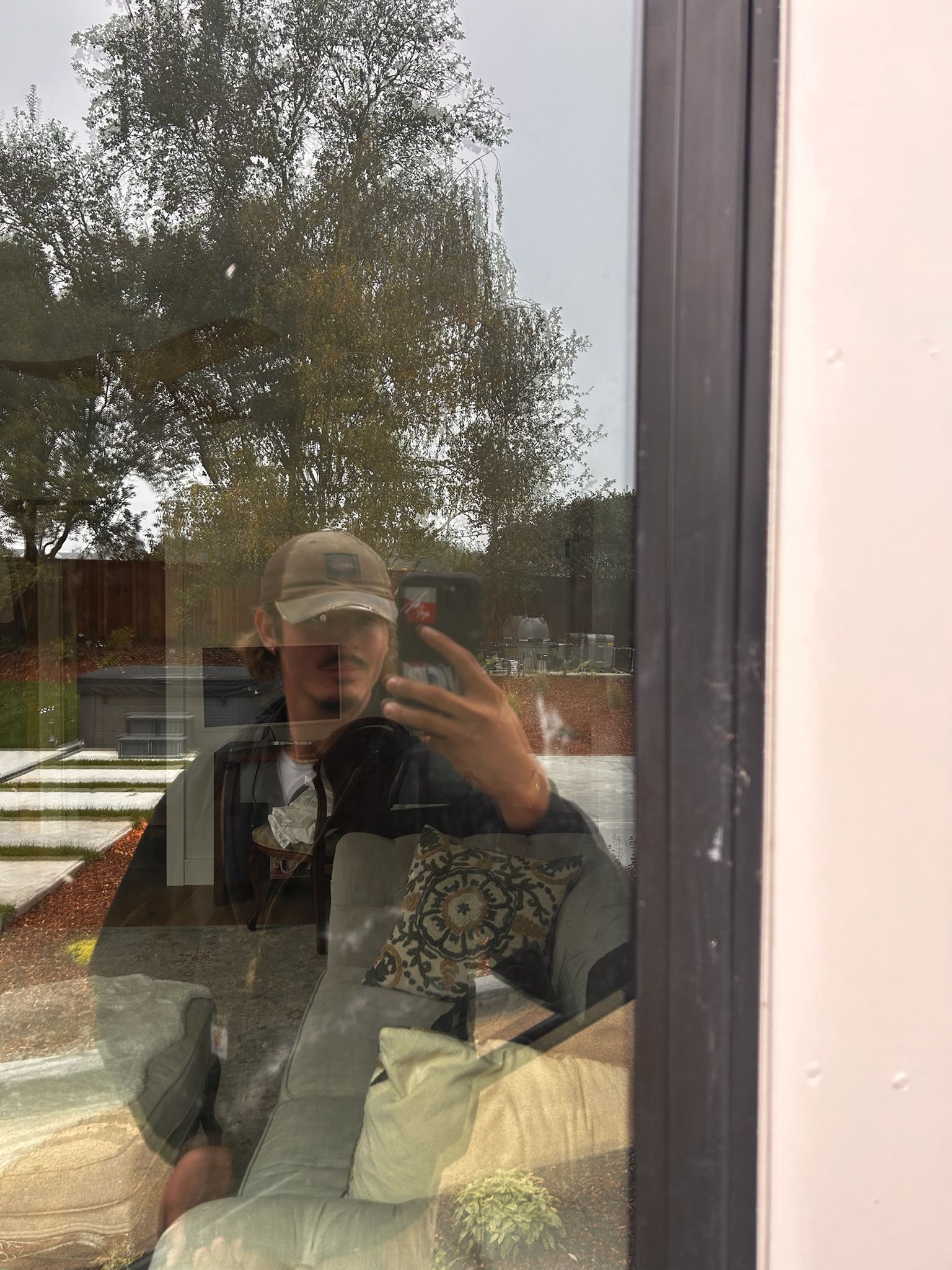 Person in a cap and jacket taking a selfie in a window, reflected trees in background.