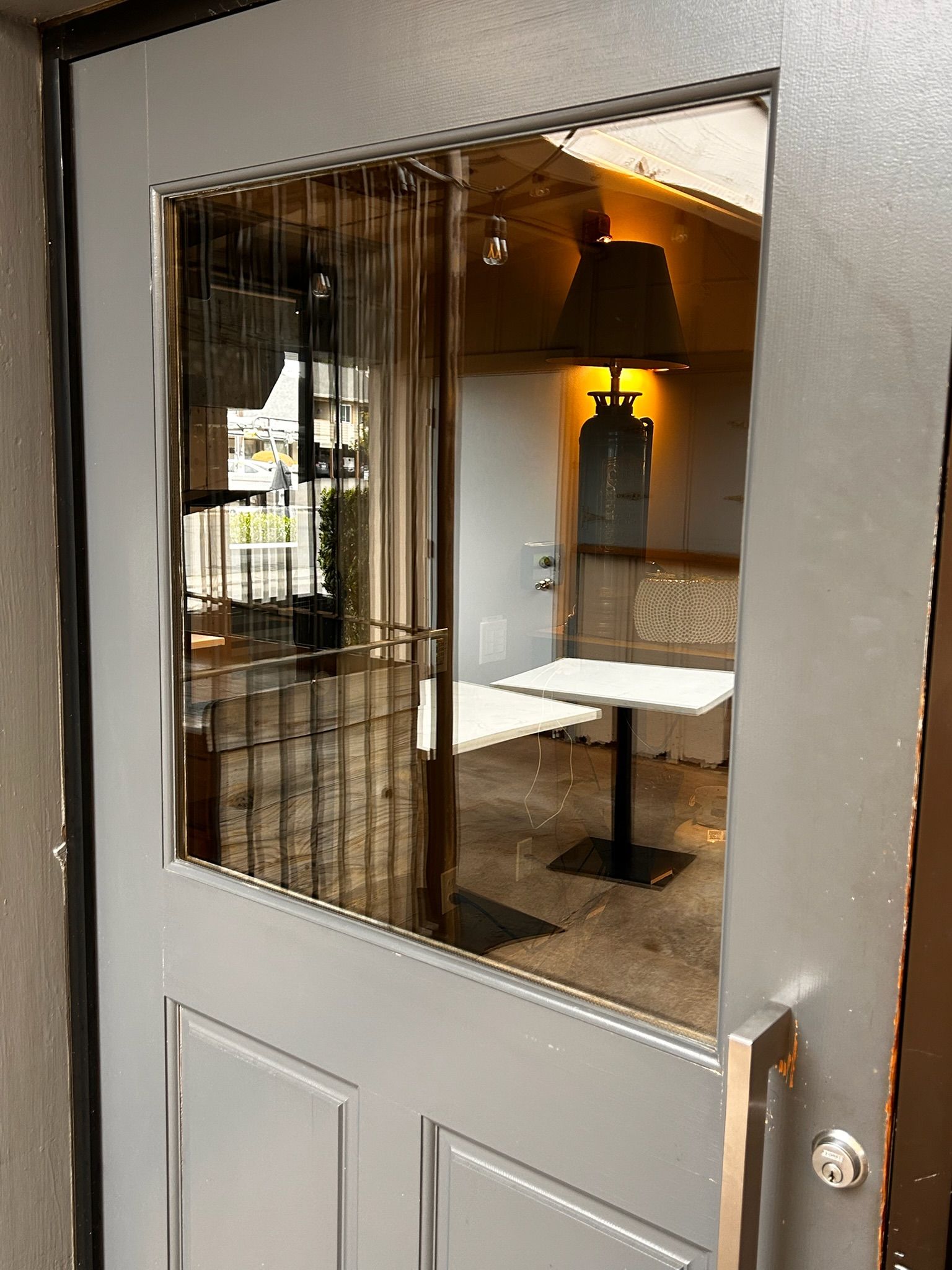 Gray door with glass panel revealing restaurant interior: tables, lamp, security bars.