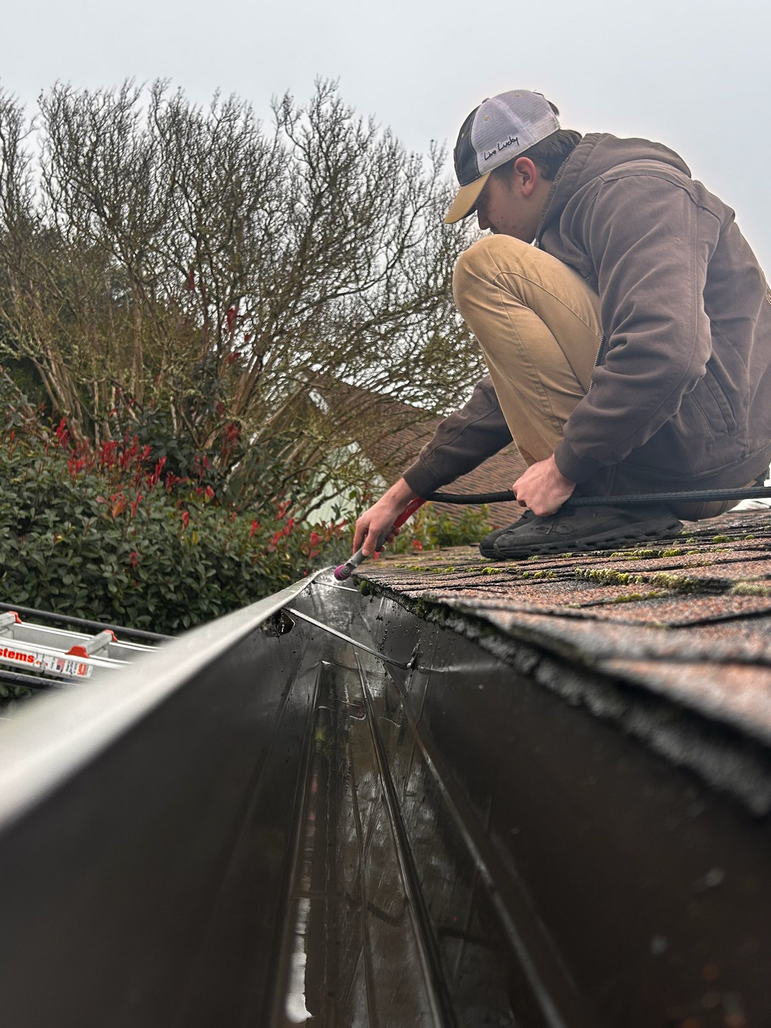 Person cleaning a gutter on a rooftop. The person wears a baseball cap and brown jacket.