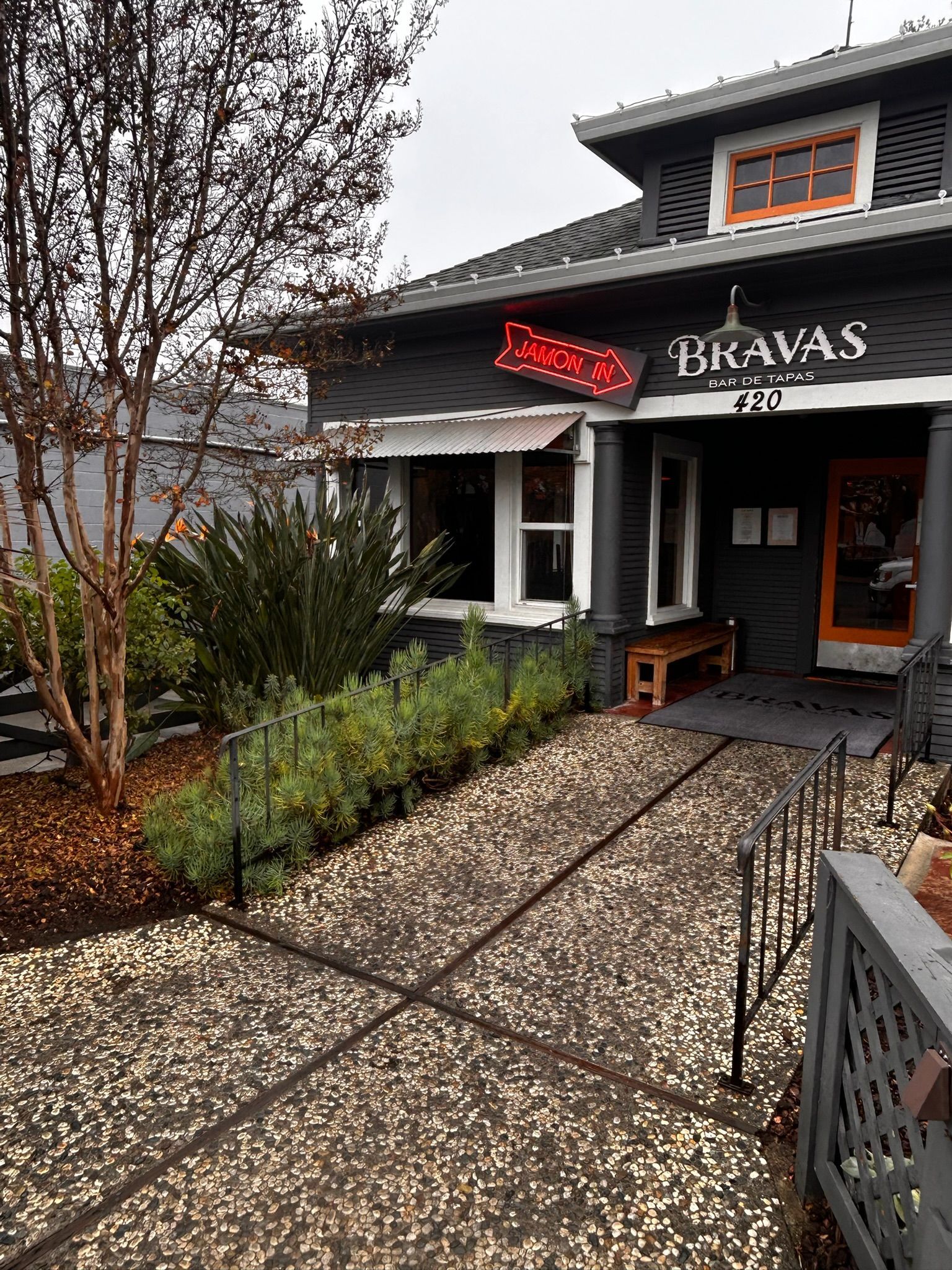 Exterior view of Bravas restaurant with dark gray facade and a sign, pathway, and landscaping.