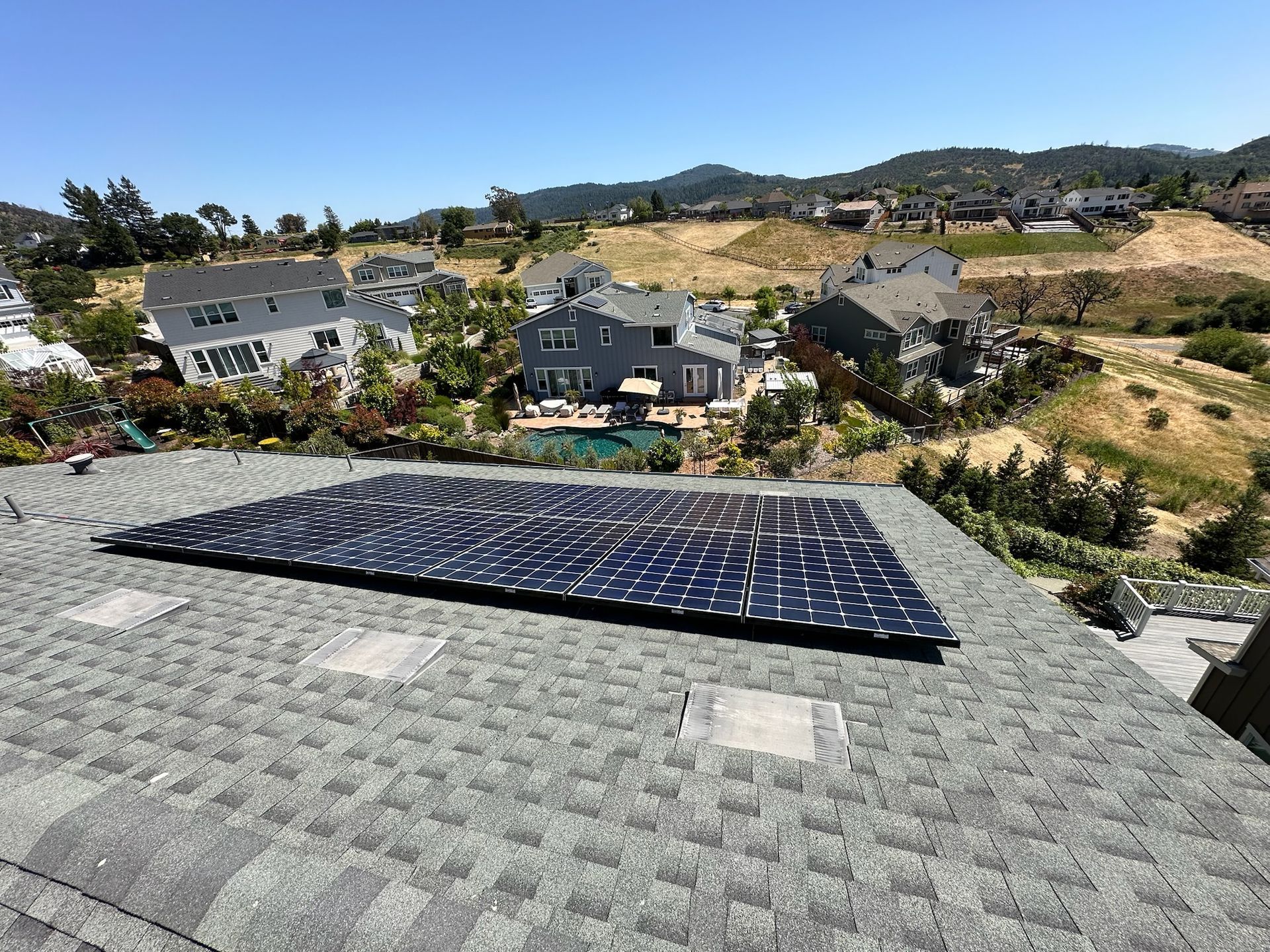Solar panels on a roof overlooking a residential area with hills and clear skies.