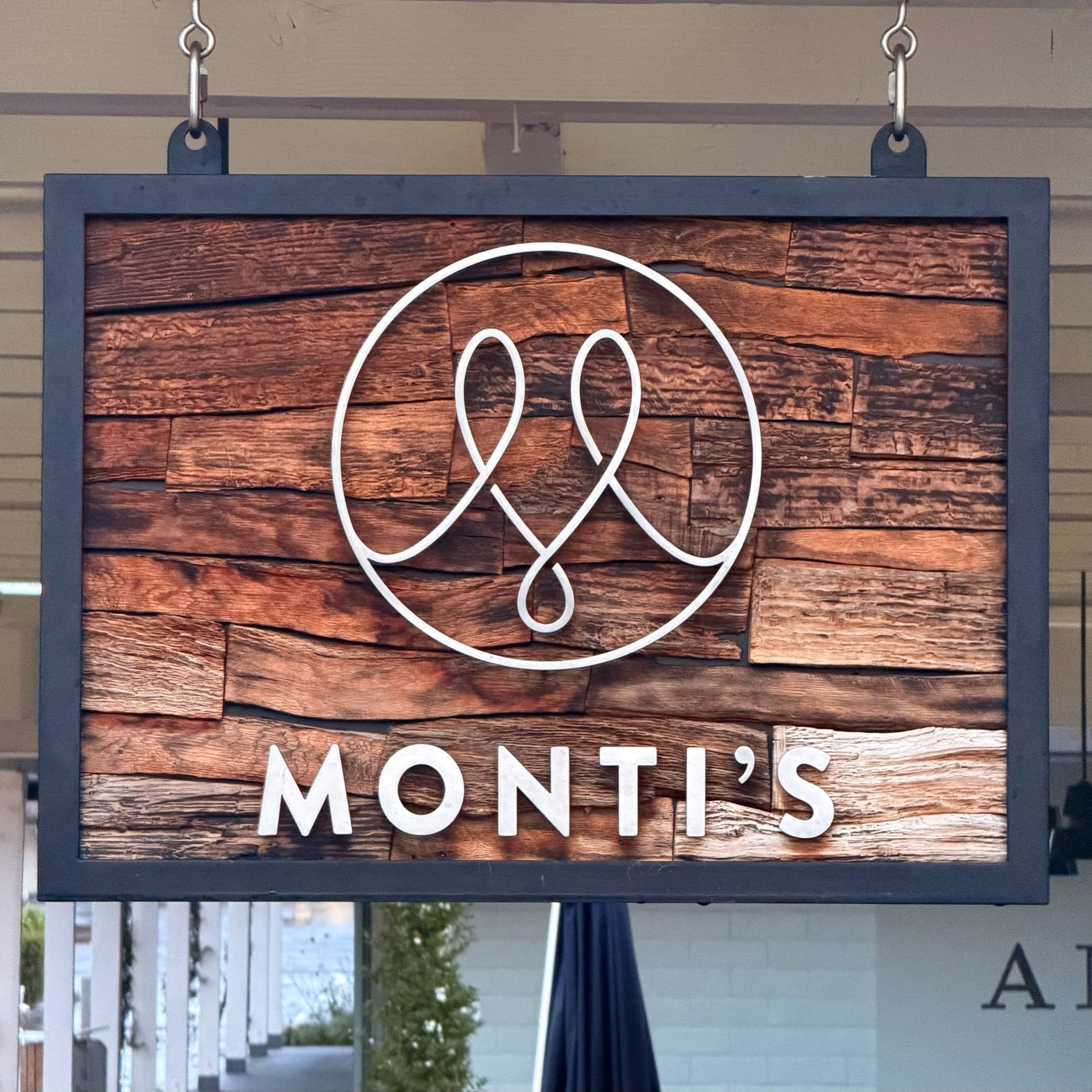 Sign for Monti's restaurant. Wooden background, white logo with intertwining lines, black frame, hanging outdoors.