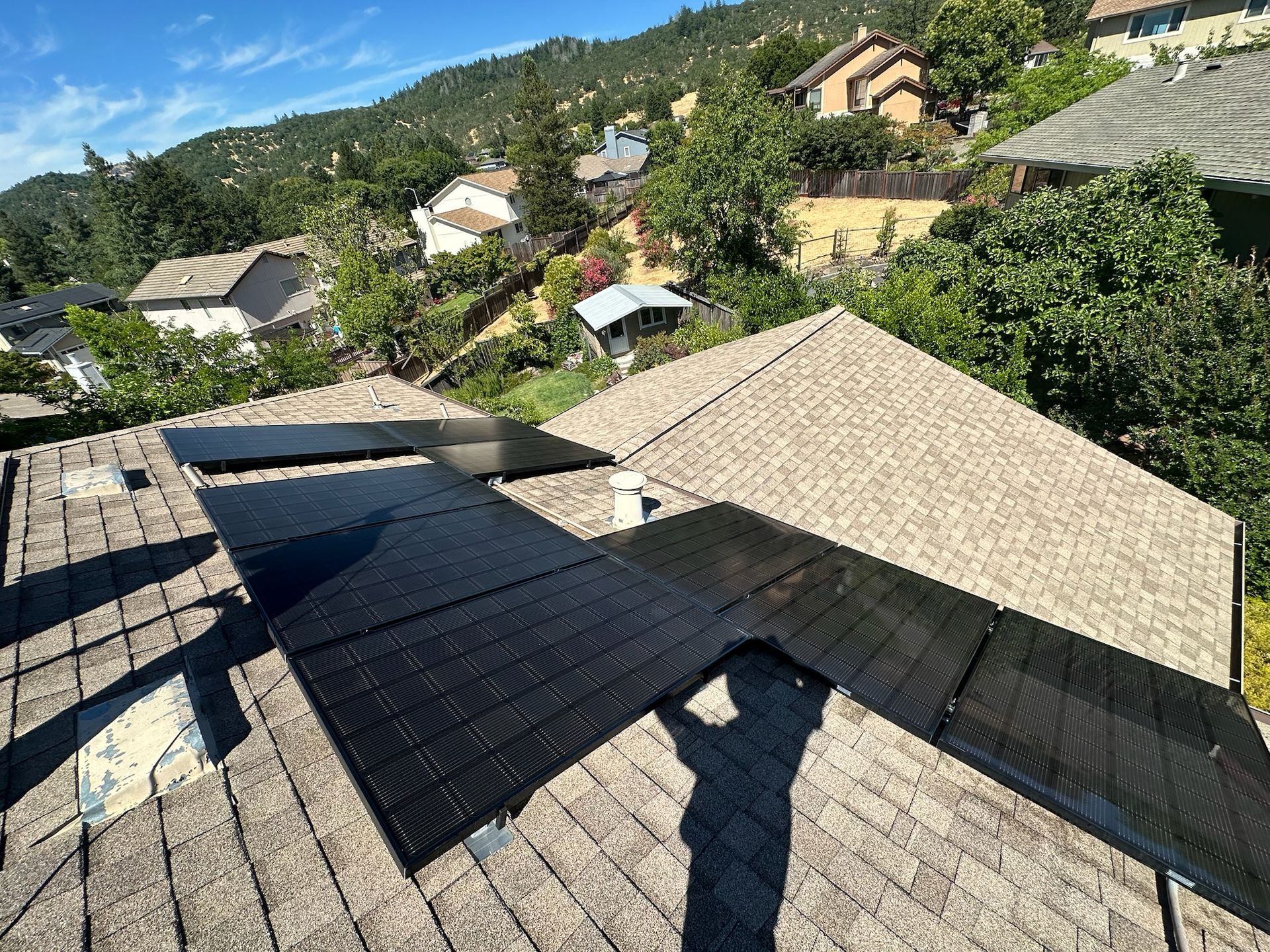 Solar panels on a brown shingled roof, set against a backdrop of trees and houses on a hillside.