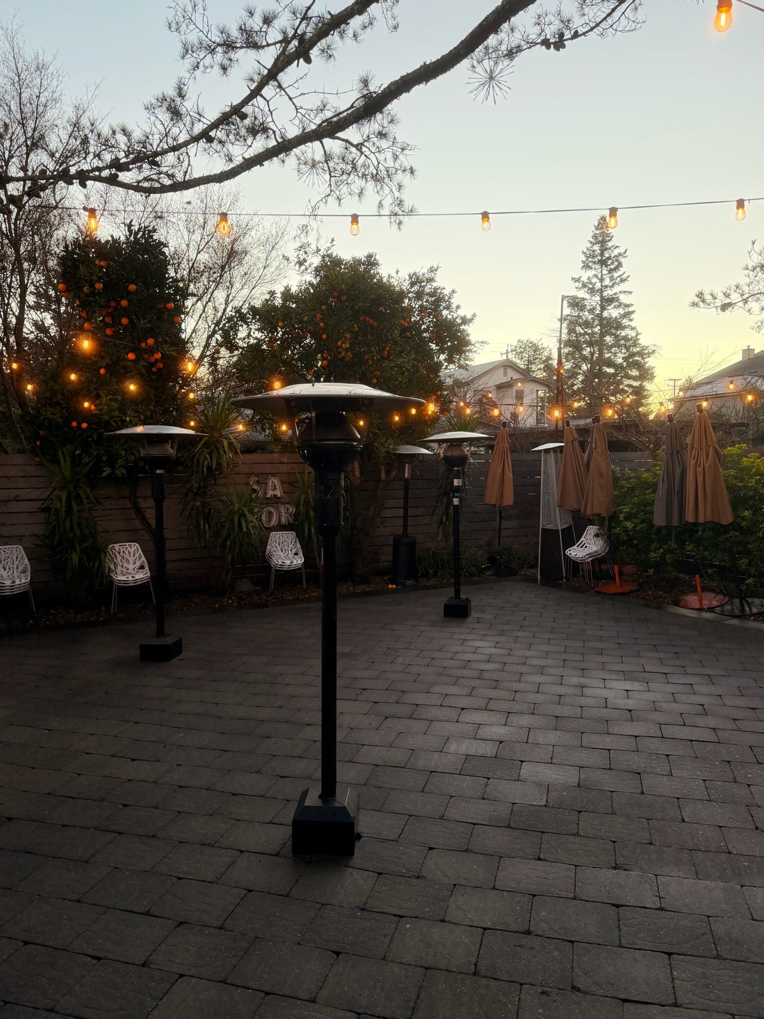 Patio at dusk with heaters, string lights, brick pavers, and trees.