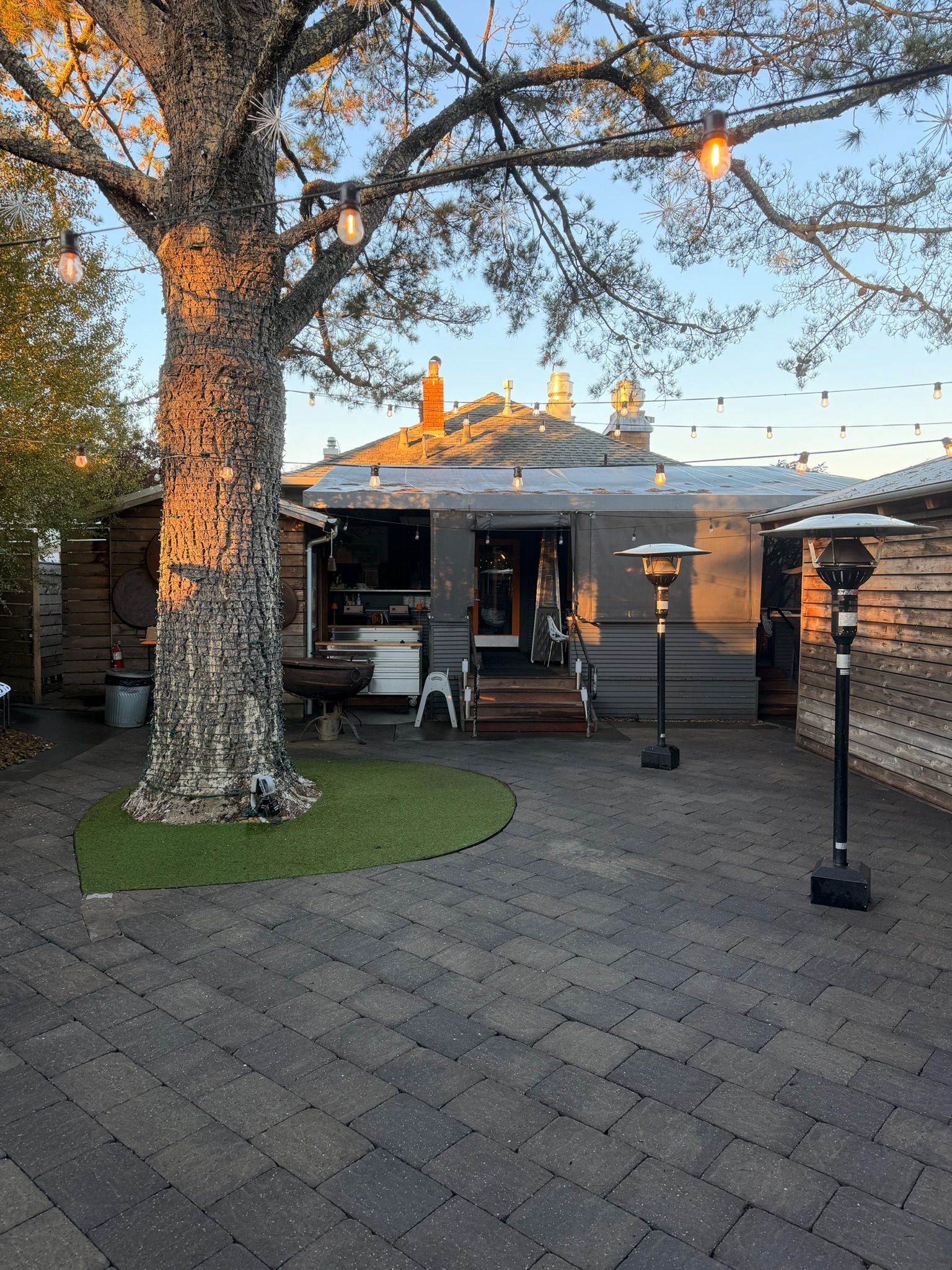 Outdoor patio with a tree, string lights, and a restaurant. Gray brick paving and space heaters.