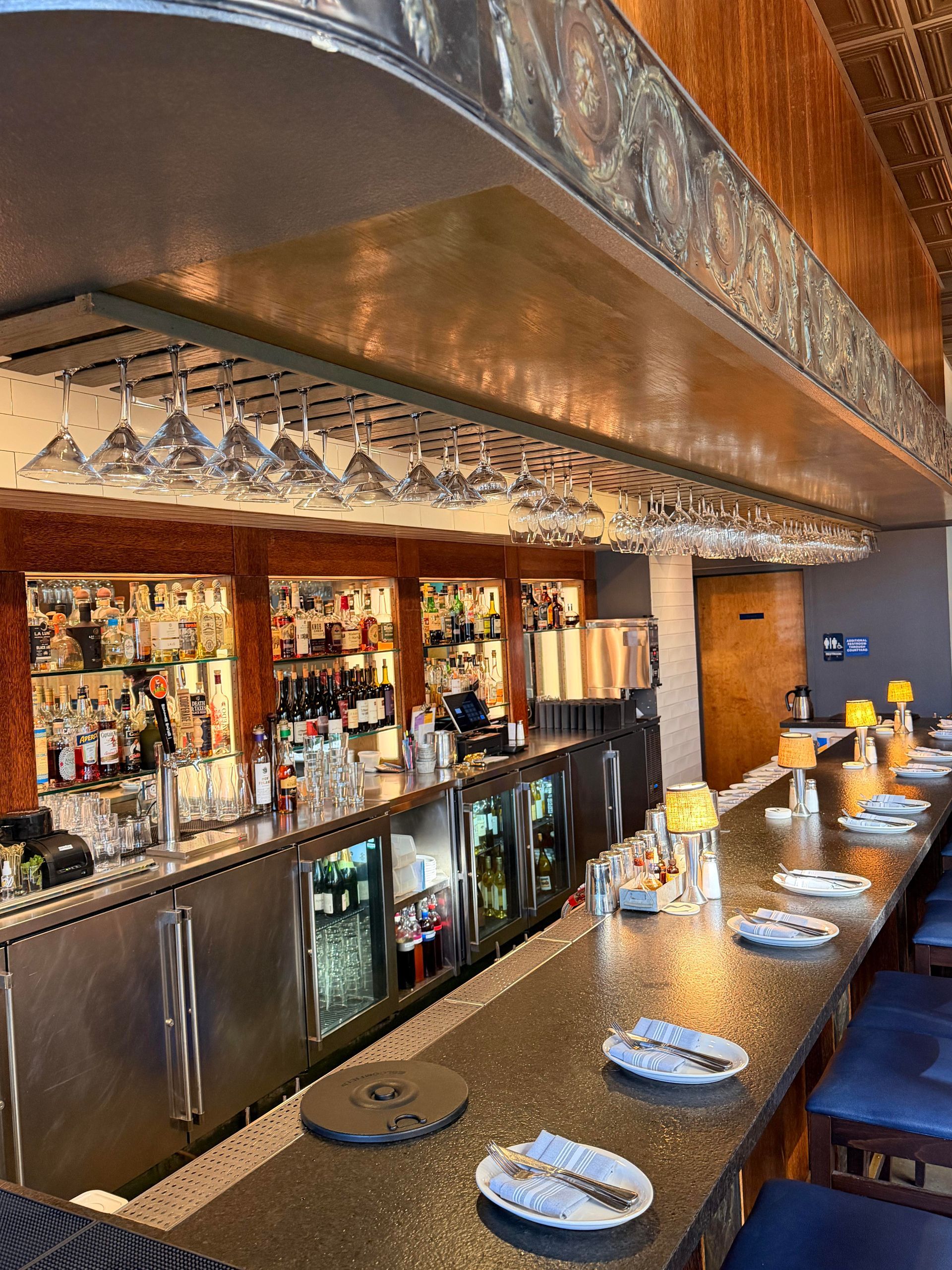 Bar interior with stainless steel bar, glassware, bottles, and blue bar stools.
