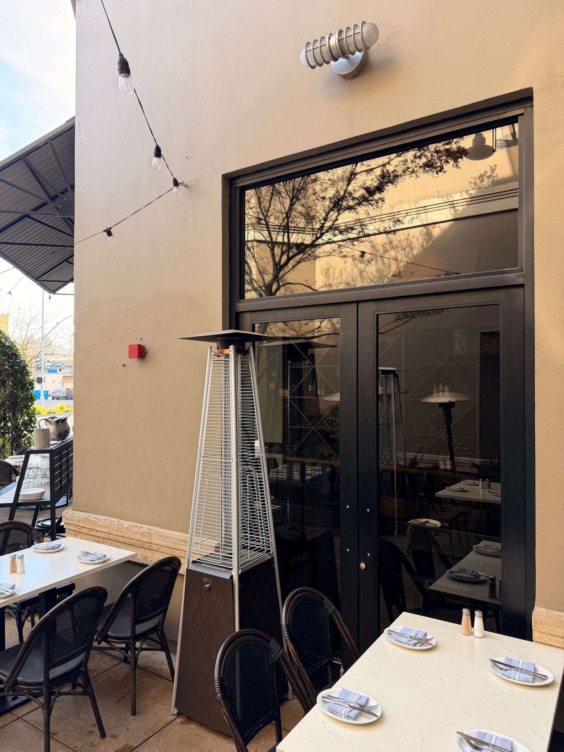 Outdoor restaurant patio with tables, chairs, heater, and dark glass doors.