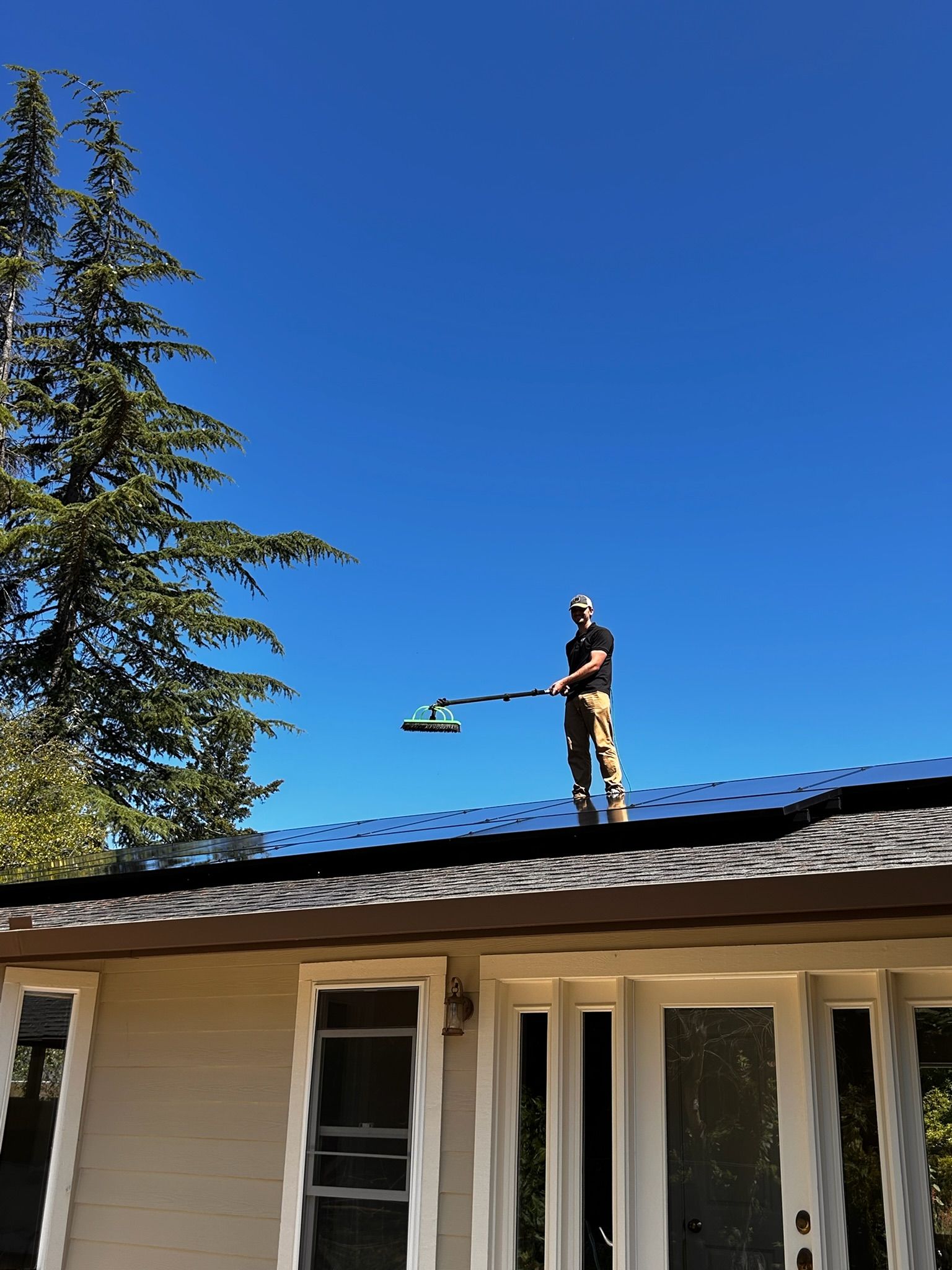Person on a roof cleaning solar panels with a long-handled brush under a blue sky.