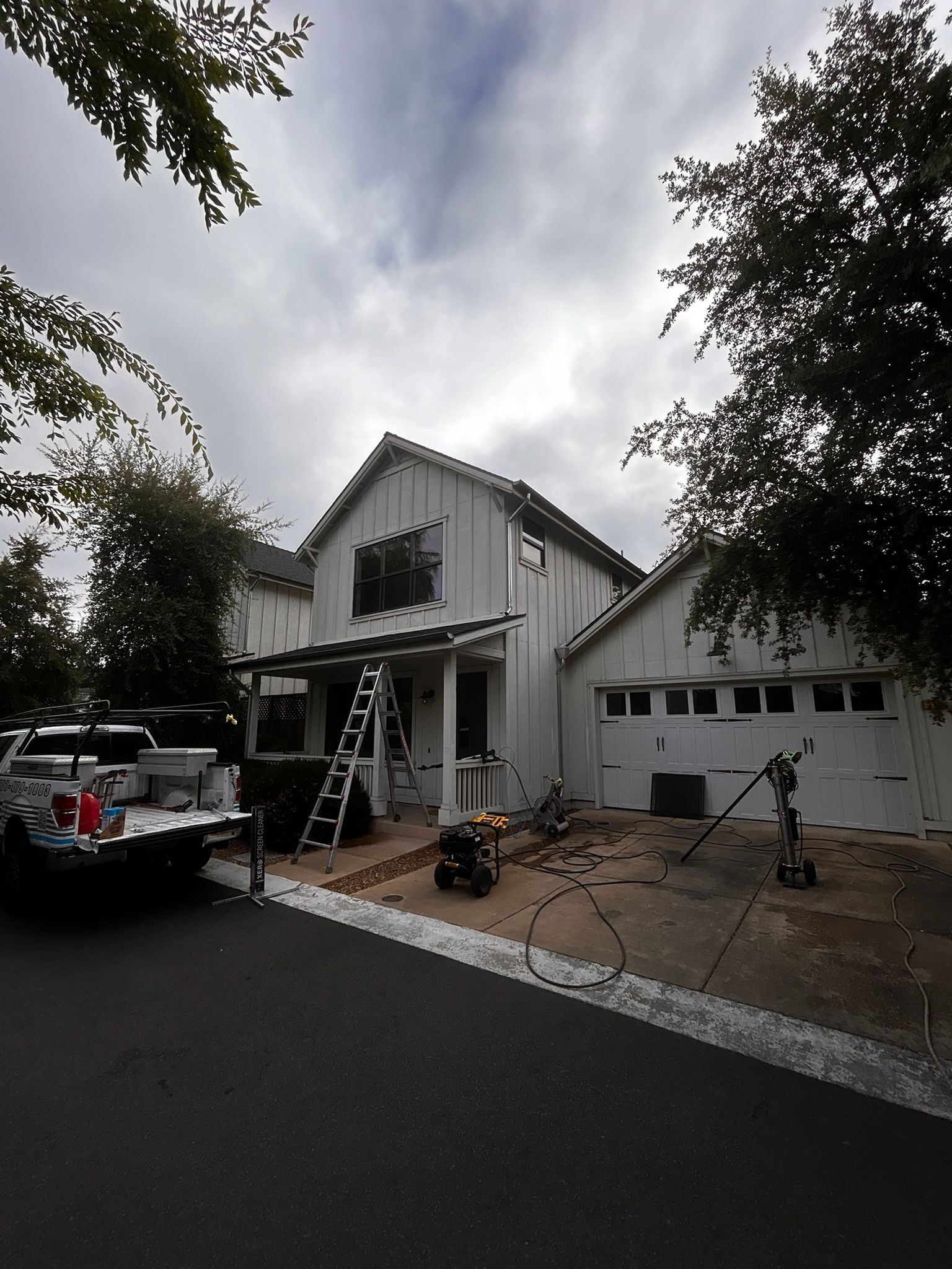 White house with ladder, tools, and a truck parked outside on a cloudy day.