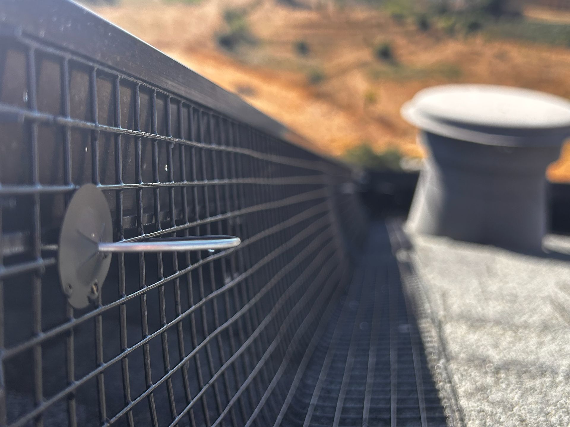 Close-up of a metal grate with a small, flat metal piece attached. A blurred vent is in the background.