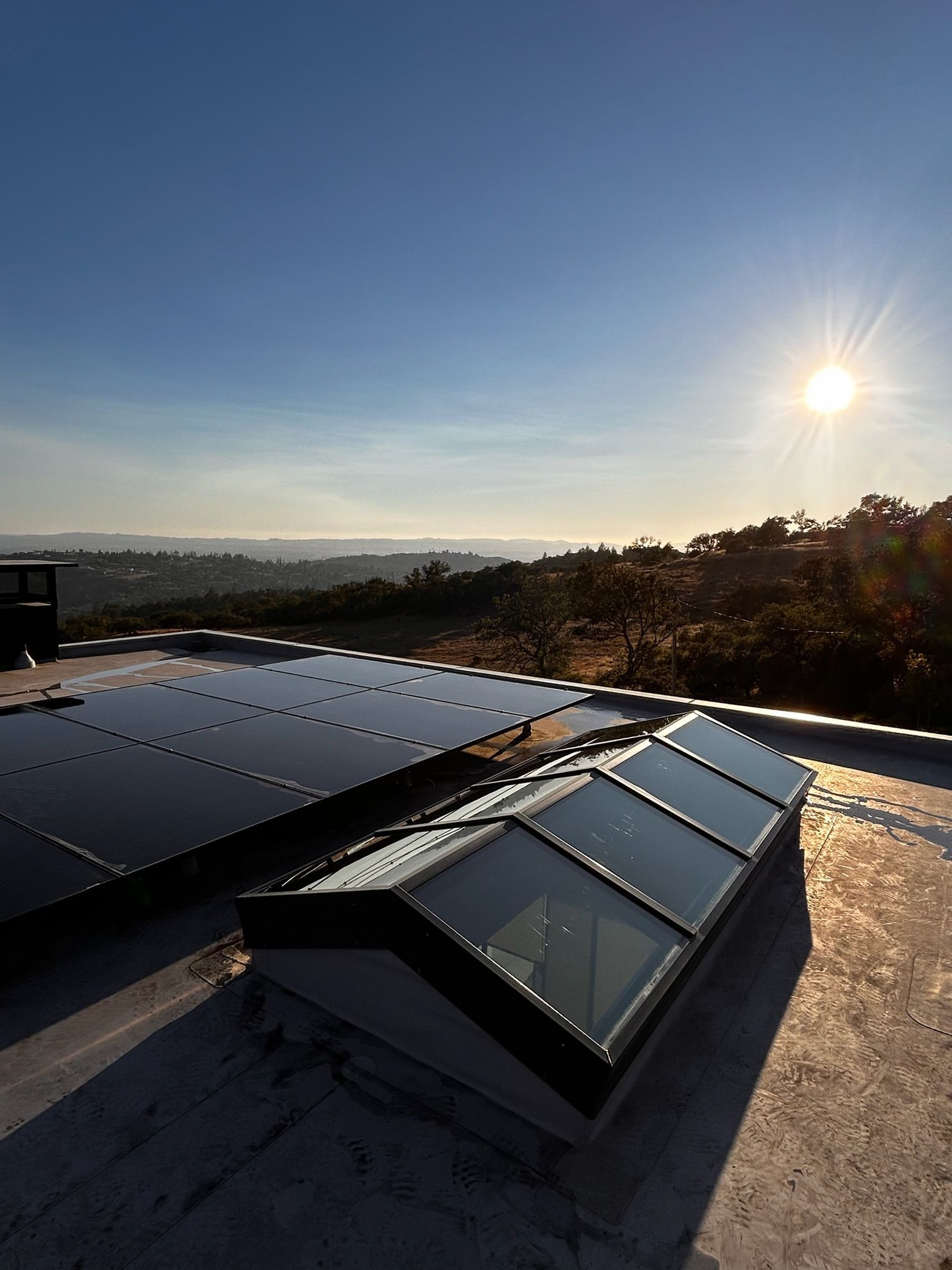 Rooftop with solar panels and a skylight under a bright sun; hilly landscape in background.