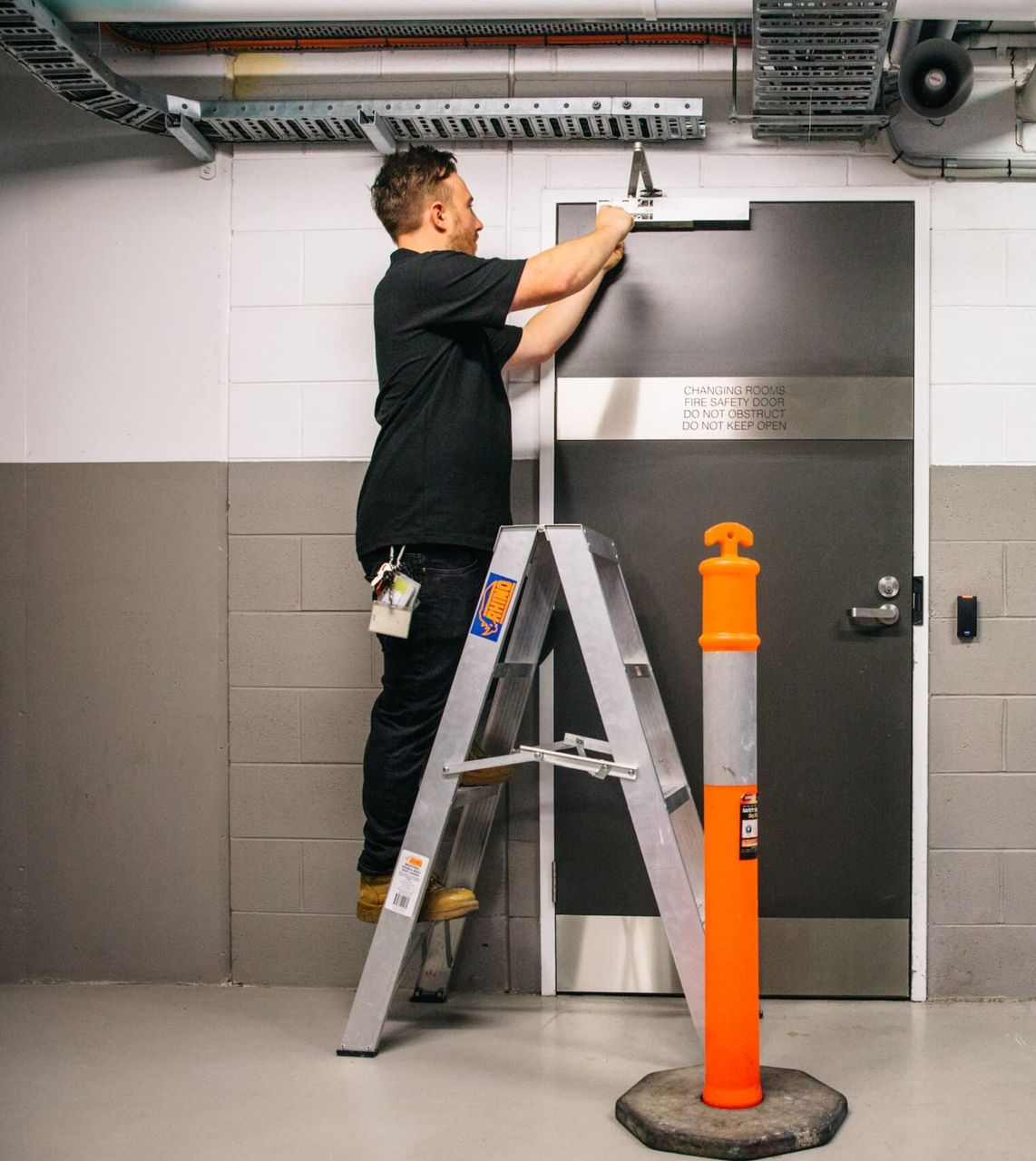 Man on a ladder installing hardware on a dark door in a commercial building. Orange bollard visible.