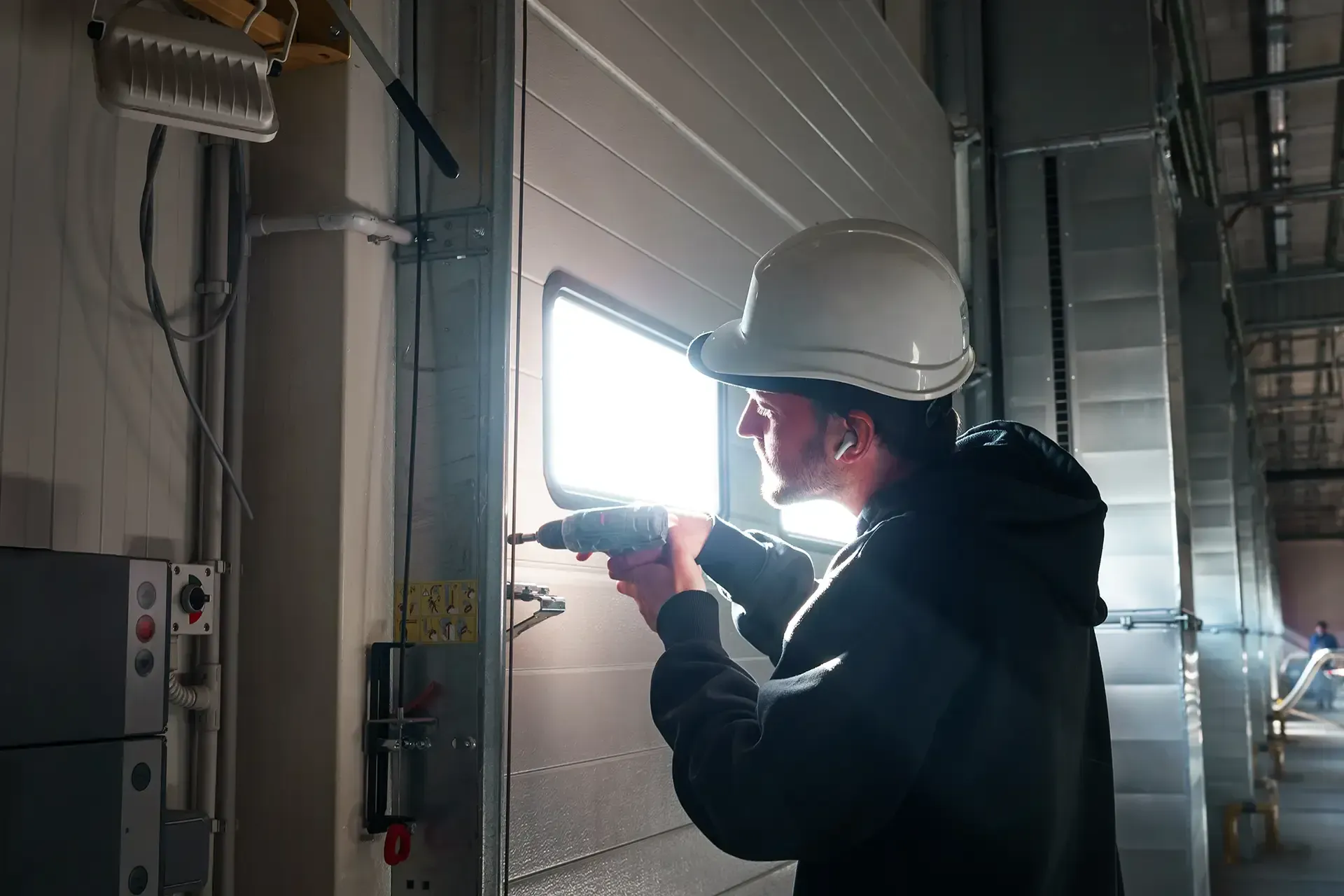 Person in hard hat using a drill on a garage door with window. Indoors, industrial setting.