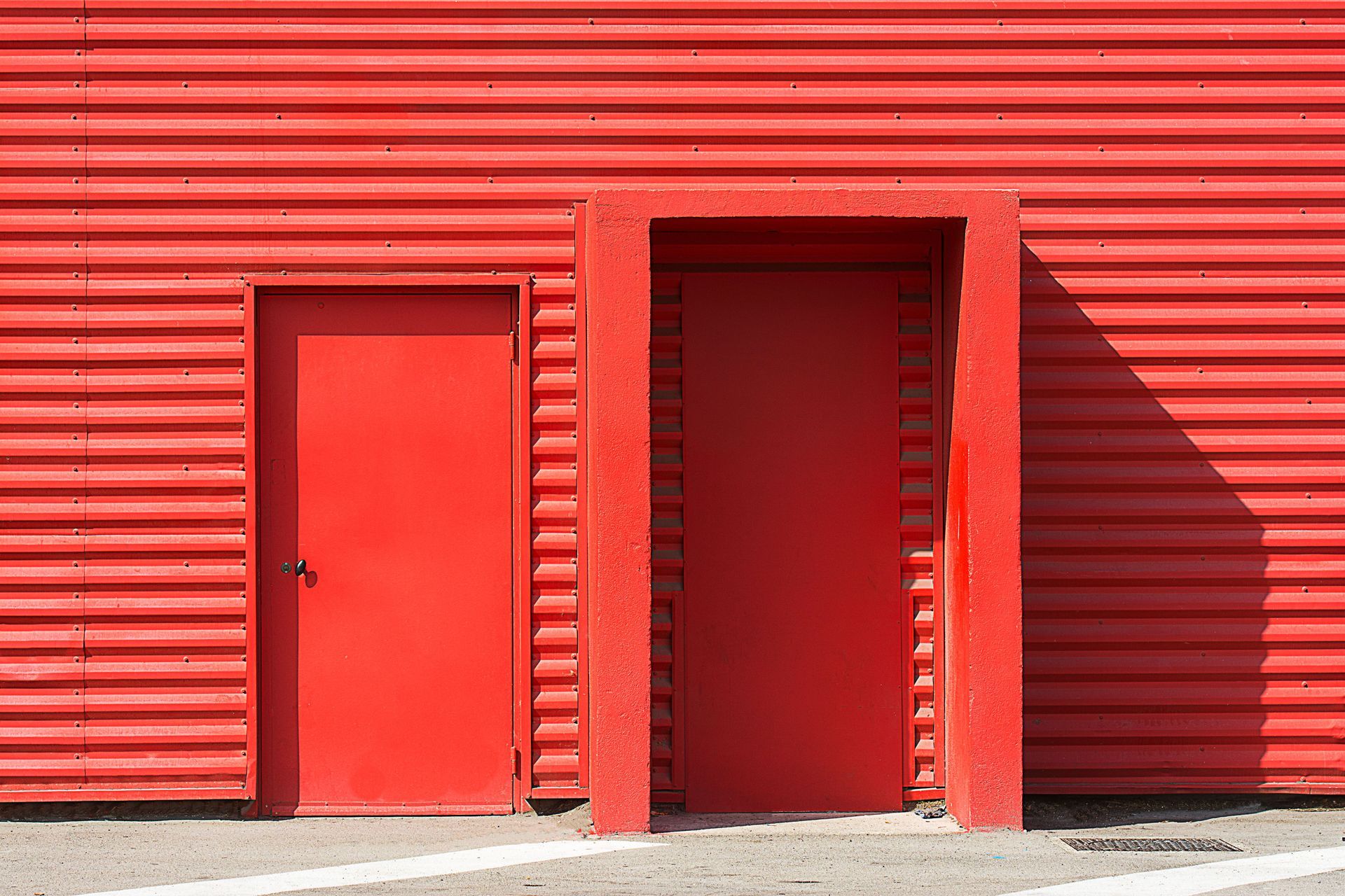 Two red doors in a red corrugated metal wall, with one narrow doorway and a shadow on the right
