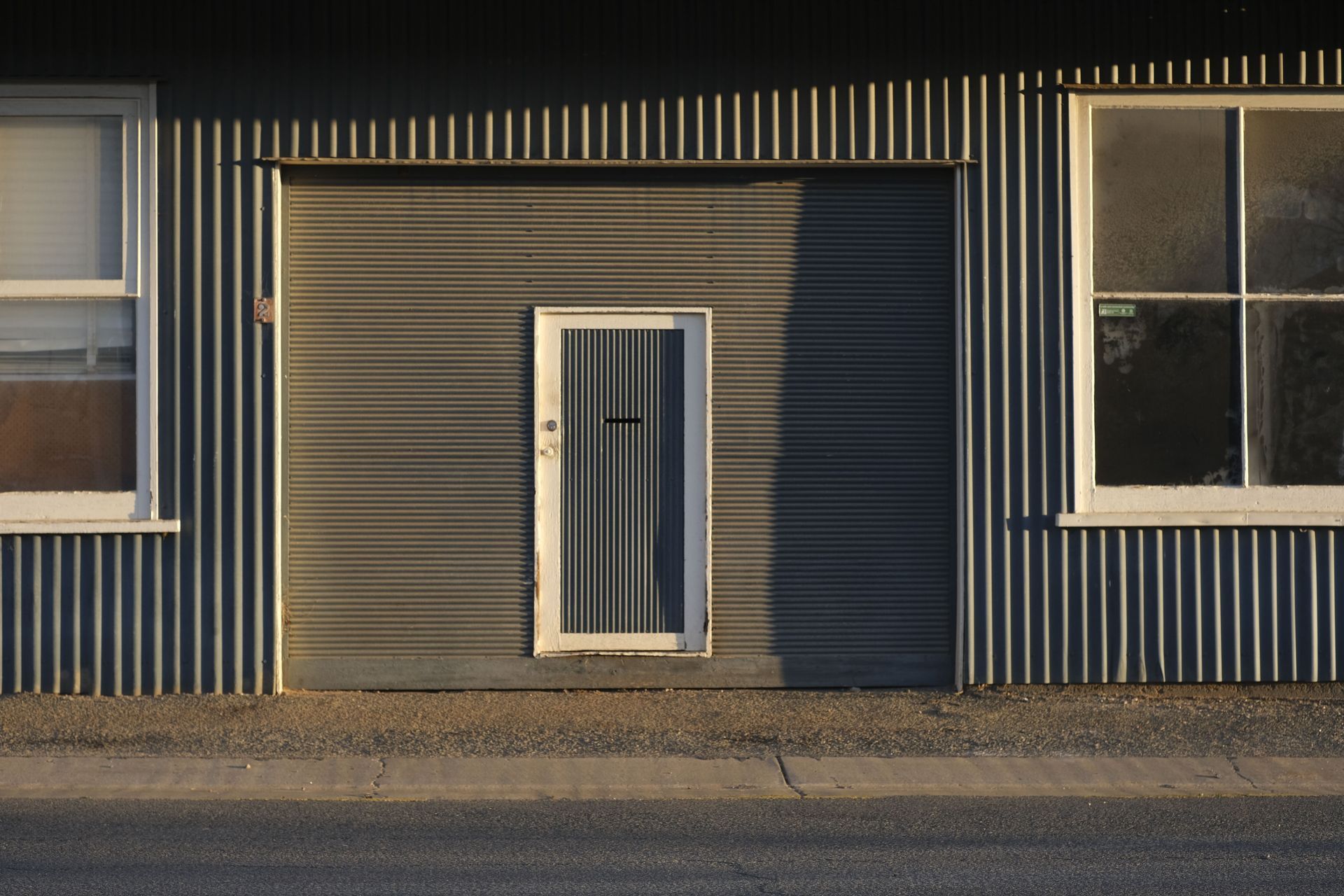 Gray corrugated building facade with two windows and a central white door by a sidewalk