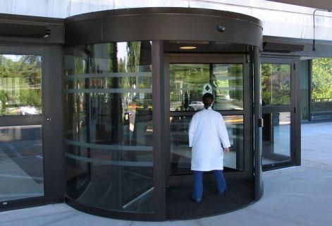 Person in white coat entering a revolving glass door at a building entrance.