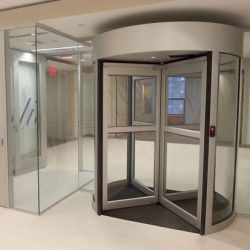 Glass revolving door in a bright interior lobby with white walls and a nearby glass entry door