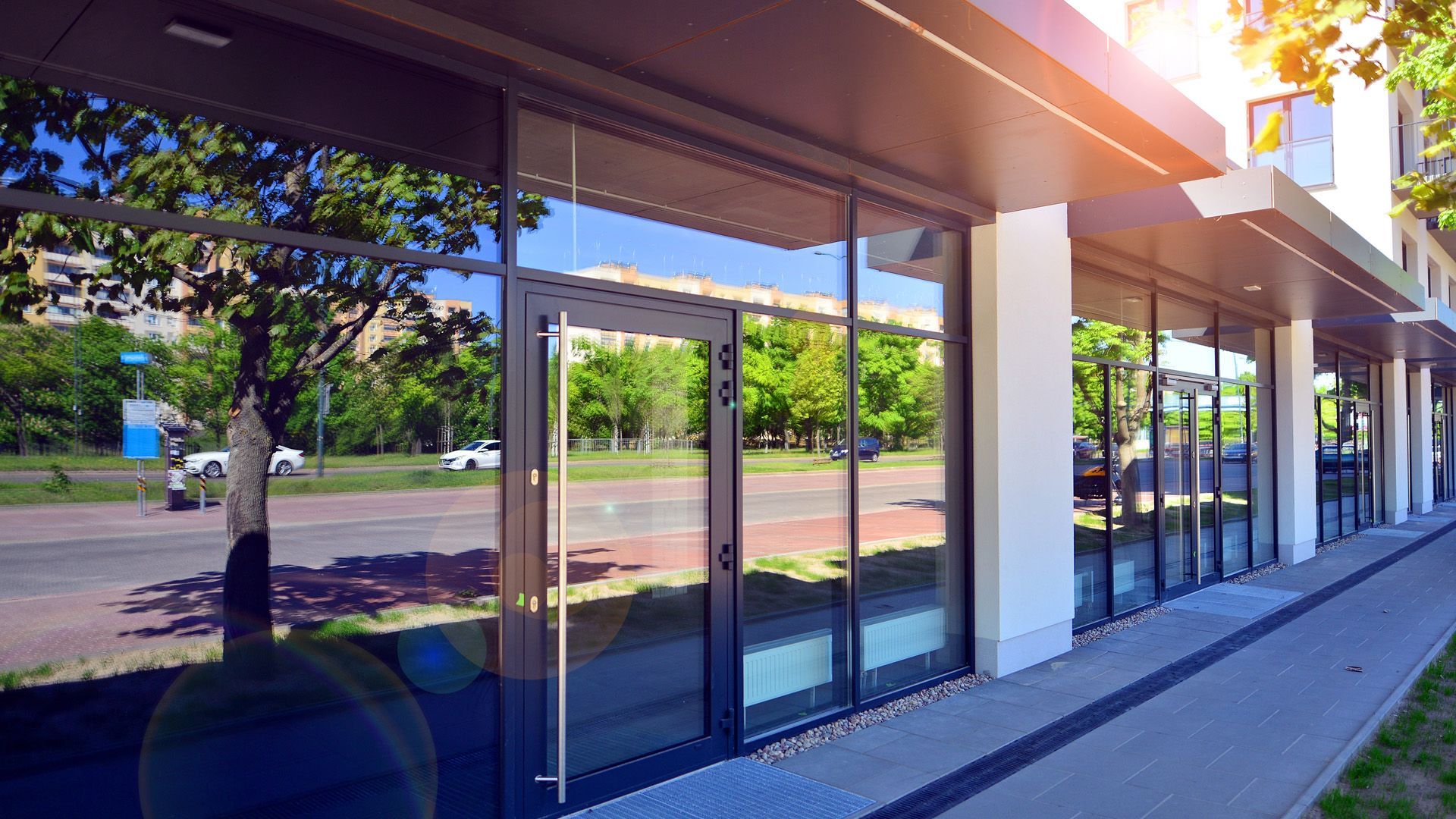 Modern commercial building with large glass windows reflecting trees and street; sunny day.