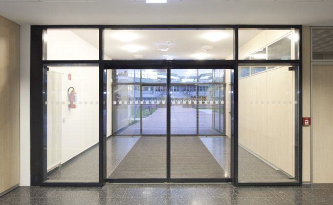 Glass automatic sliding doors lead to an outdoor walkway, flanked by white walls and a fire extinguisher.