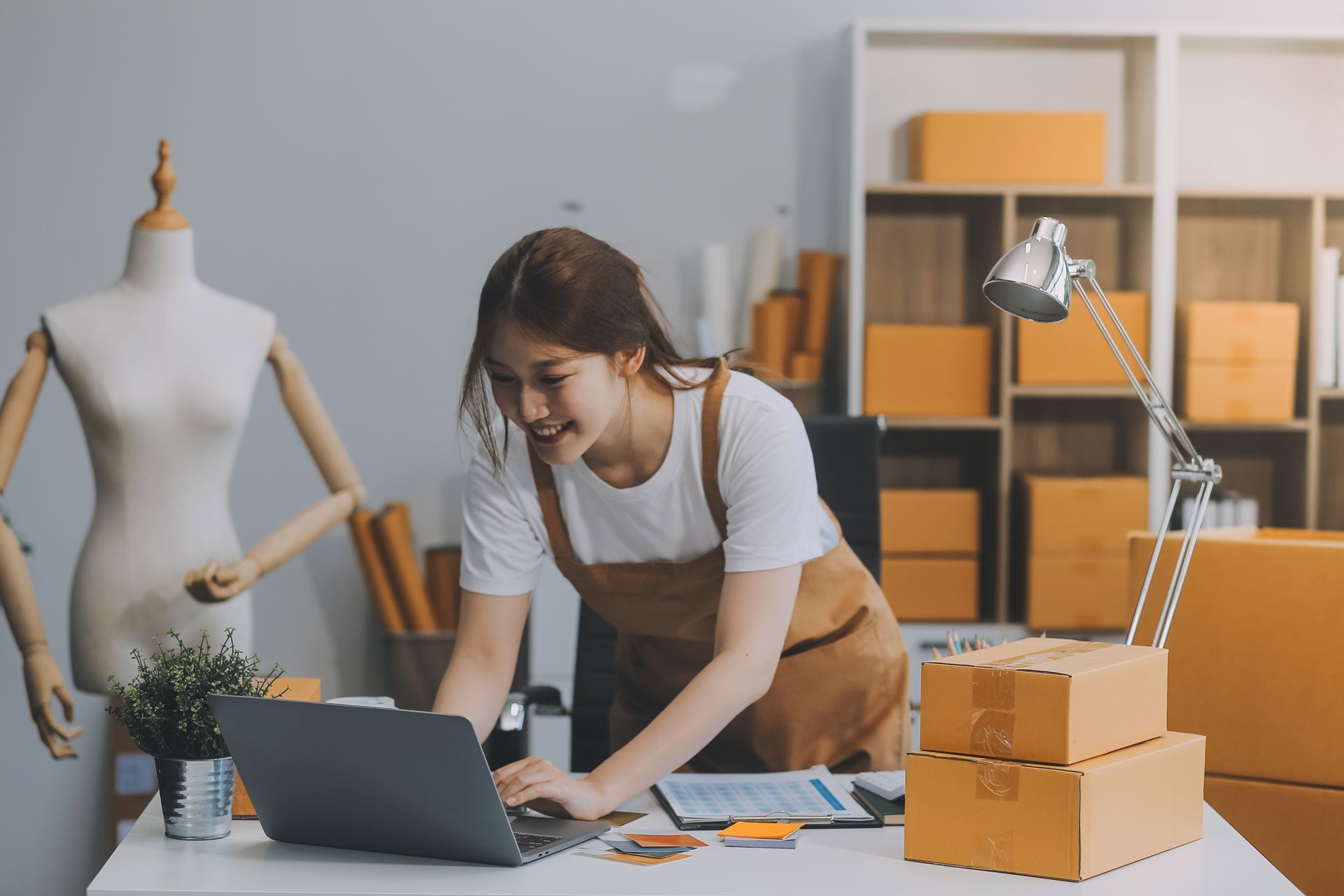 Woman working on a laptop at a desk, surrounded by boxes, mannequin, and shelves.