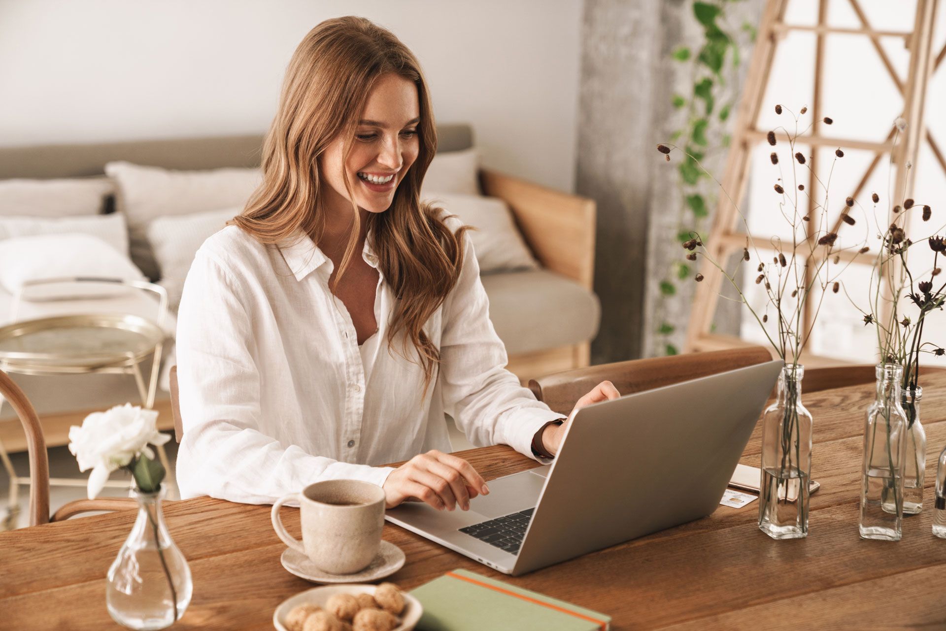 Woman smiles while typing on a laptop at a wooden table. Coffee, cookies, and a vase are present.
