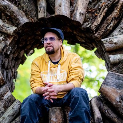 Ein Mann mit einer Brille und einem gelben Pulli sitzt in einem aus Holz geformten Kreis